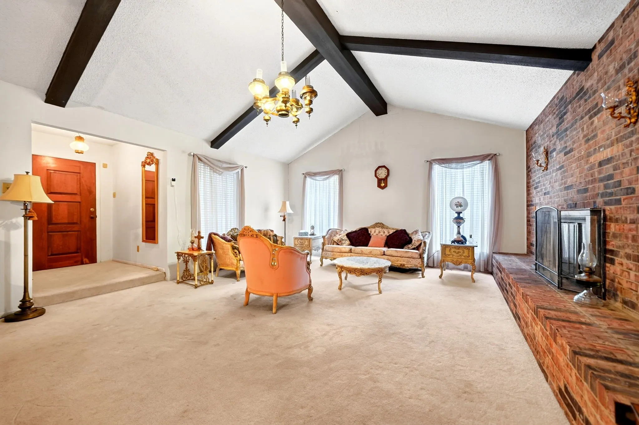 Vaulted ceiling with exposed wood beams, brick accent wall with a fireplace, large windows with drapes, and a raised entry area