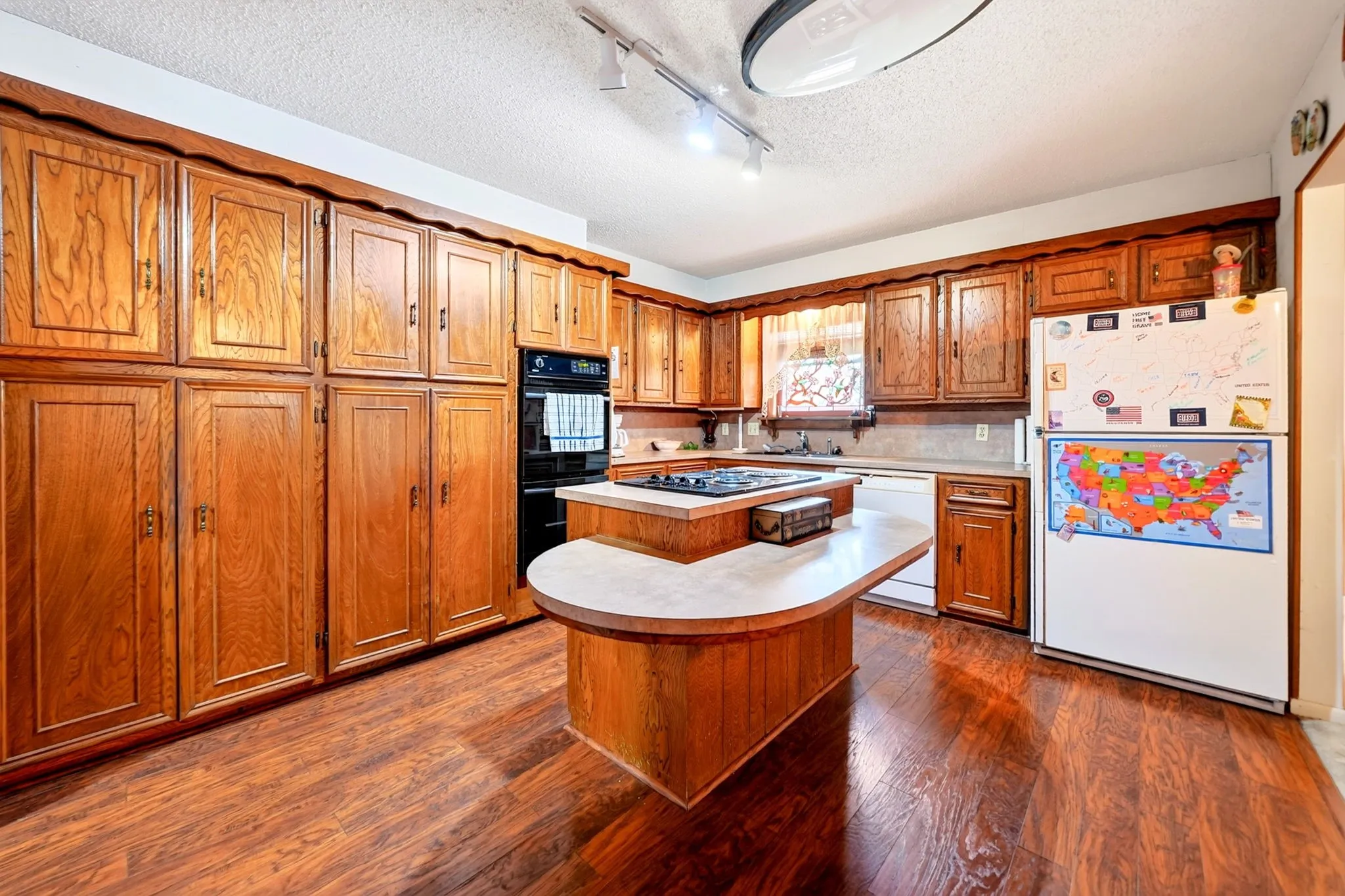 Extensive wood cabinetry, central island with integrated cooktop, double wall oven, wood-finish flooring, and track lighting