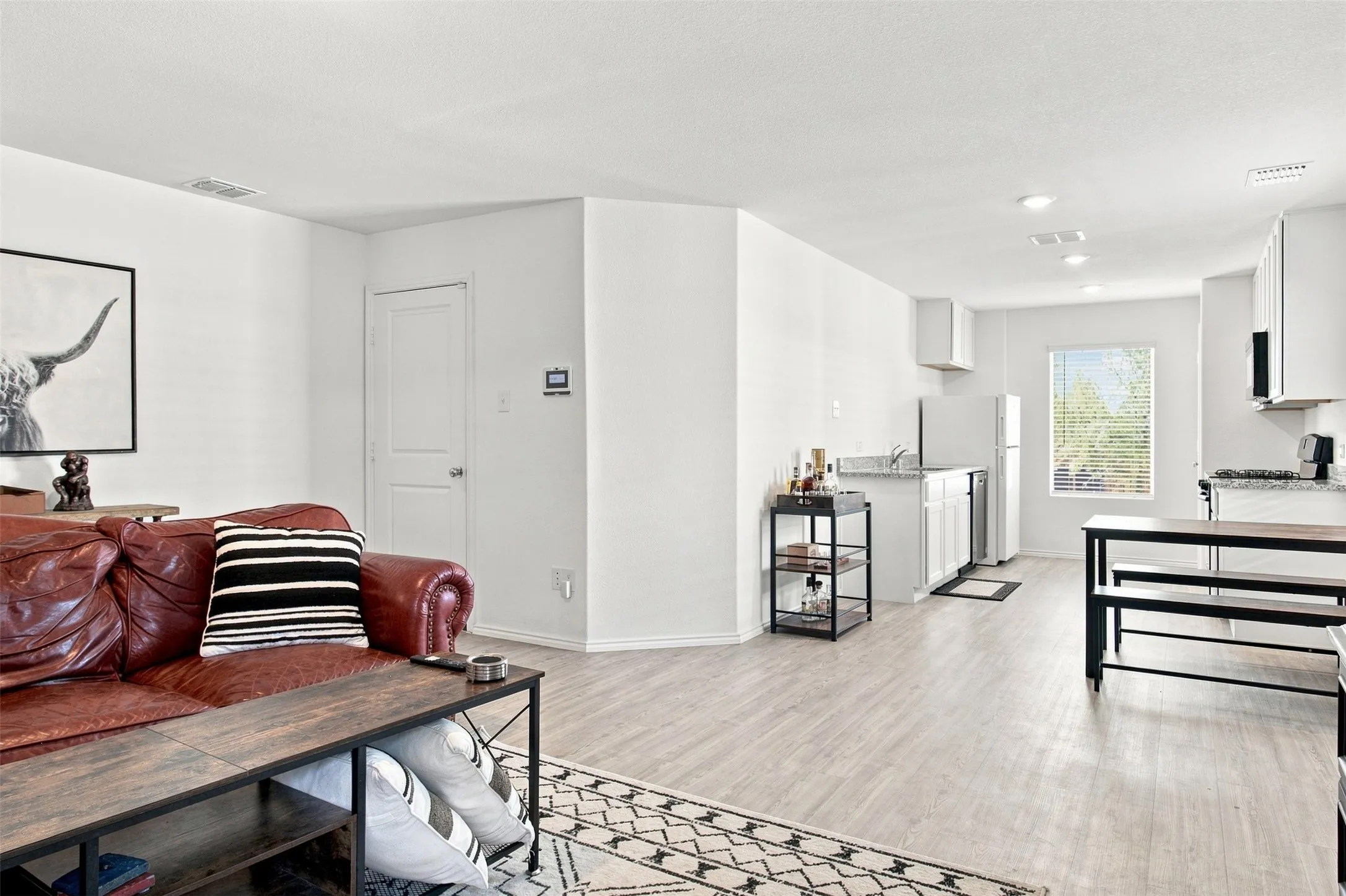 Open-concept living area featuring light wood-finish flooring, white walls, recessed lighting, and a kitchen with white cabinetry and granite countertops