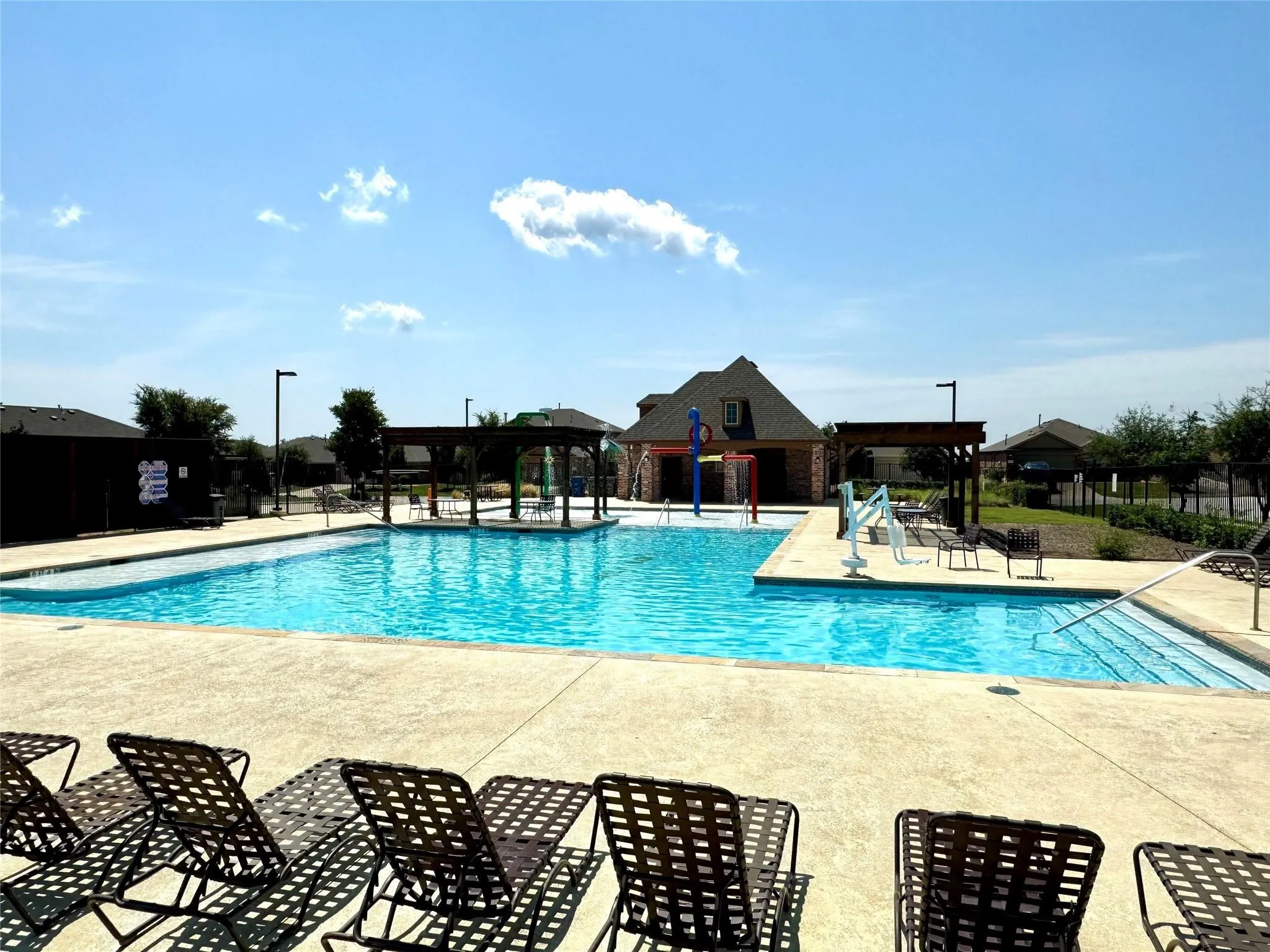Resort-style community pool featuring crystal-clear water, expansive concrete decking with lounge seating, a shallow children's play area, and a clubhouse structure with pitched roofline