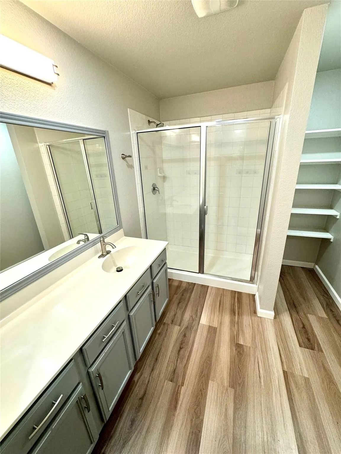 Spa-inspired primary bathroom featuring a wood-finish vanity with dual sinks, quartz countertop, framed mirror, and a frameless glass-enclosed shower with tile surround