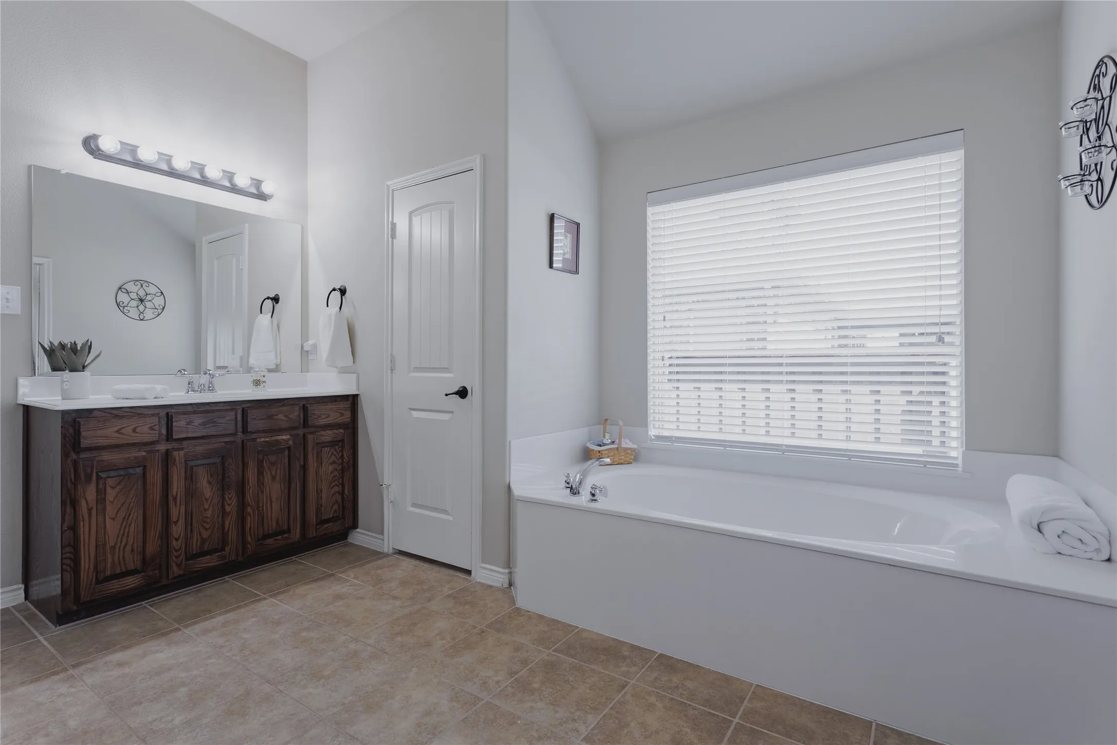 Spa-inspired primary bathroom featuring a dual-sink vanity with wood-finish cabinetry and quartz countertop, a freestanding soaking tub positioned beneath a large picture window with plantation shutters, and neutral stone-finish flooring throughout