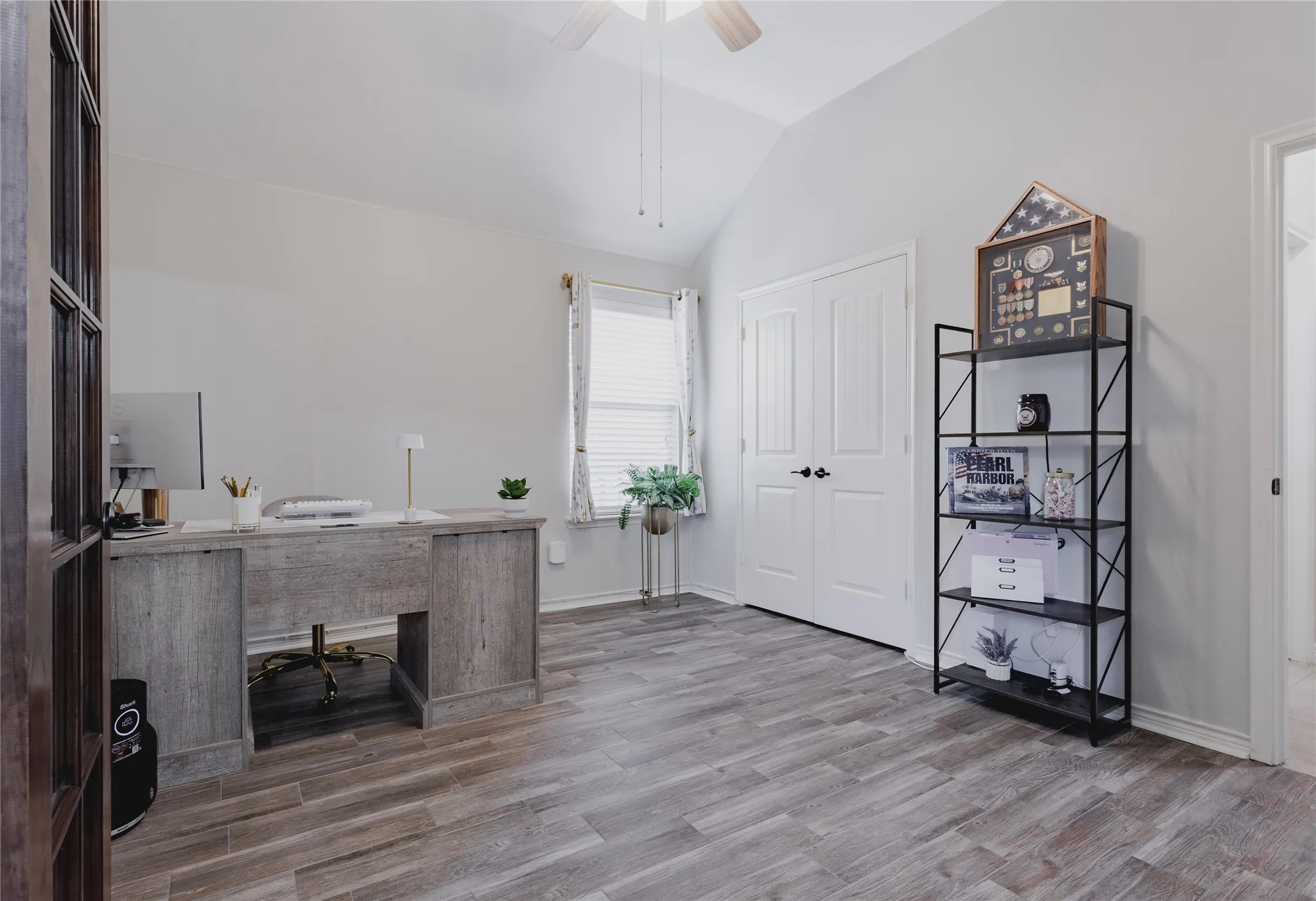 Bright home office featuring wood-finish flooring, a built-in desk with storage, dual white panel doors, a metal shelving unit with five tiers, and a large window providing natural light