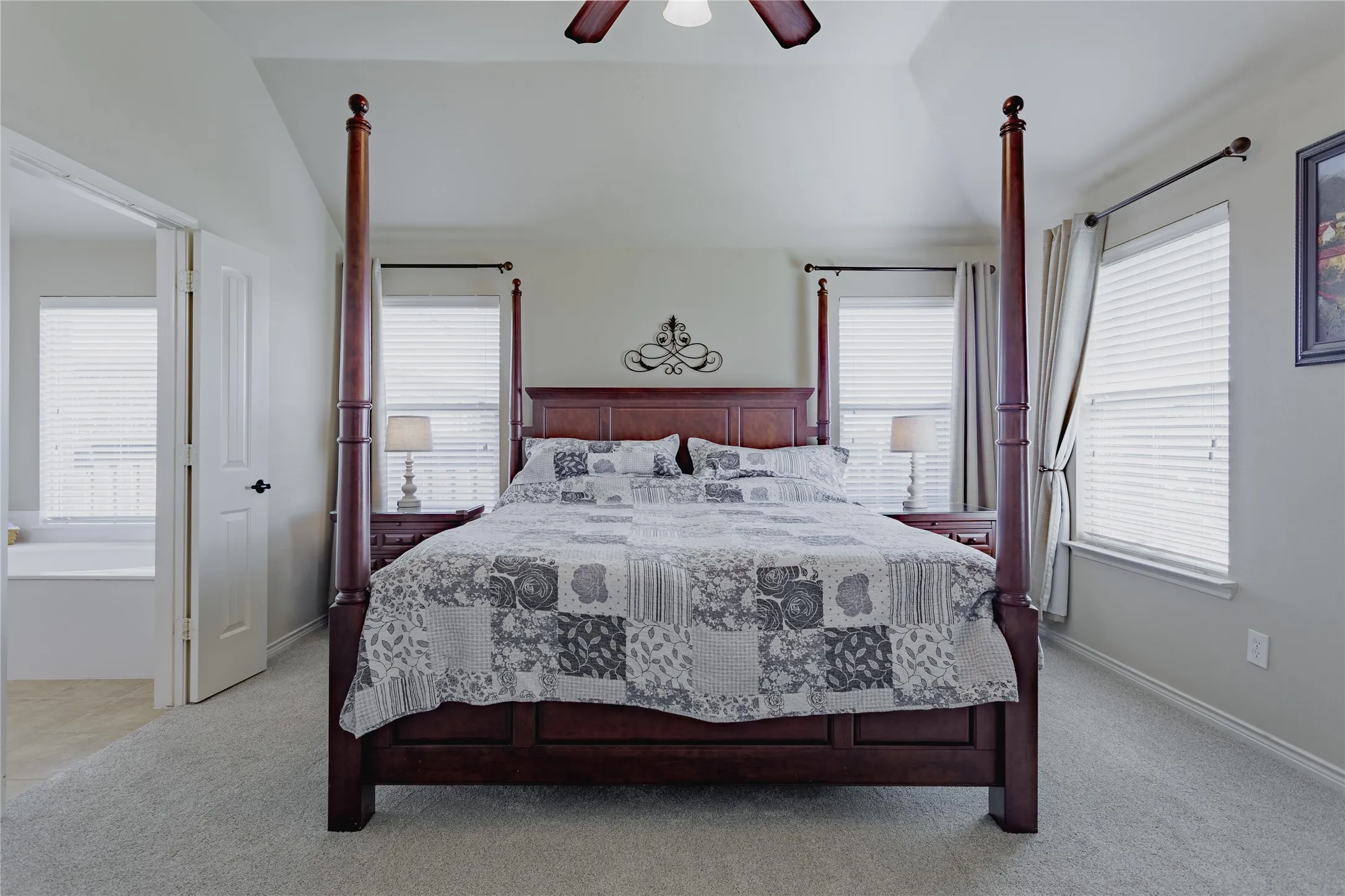 Carpeted bedroom with a vaulted ceiling, multiple windows with blinds, and an adjacent bathroom with a built-in tub