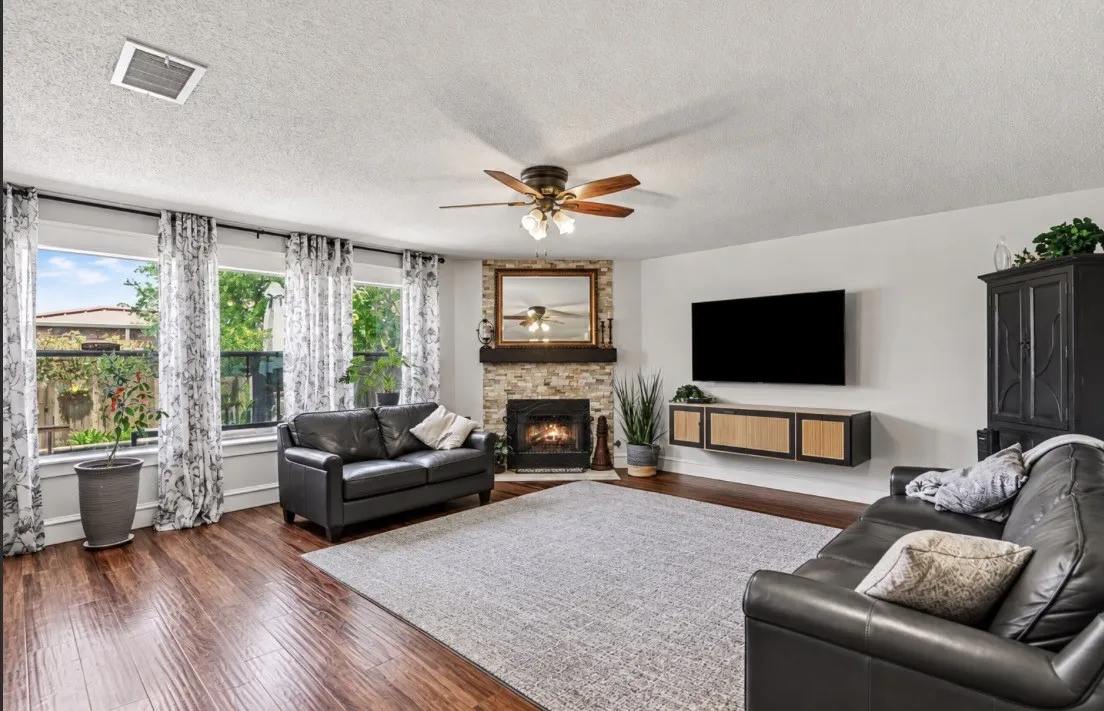 Wood-finish flooring, stone-clad fireplace with wood mantle, multiple windows, ceiling fan, and a built-in media console
