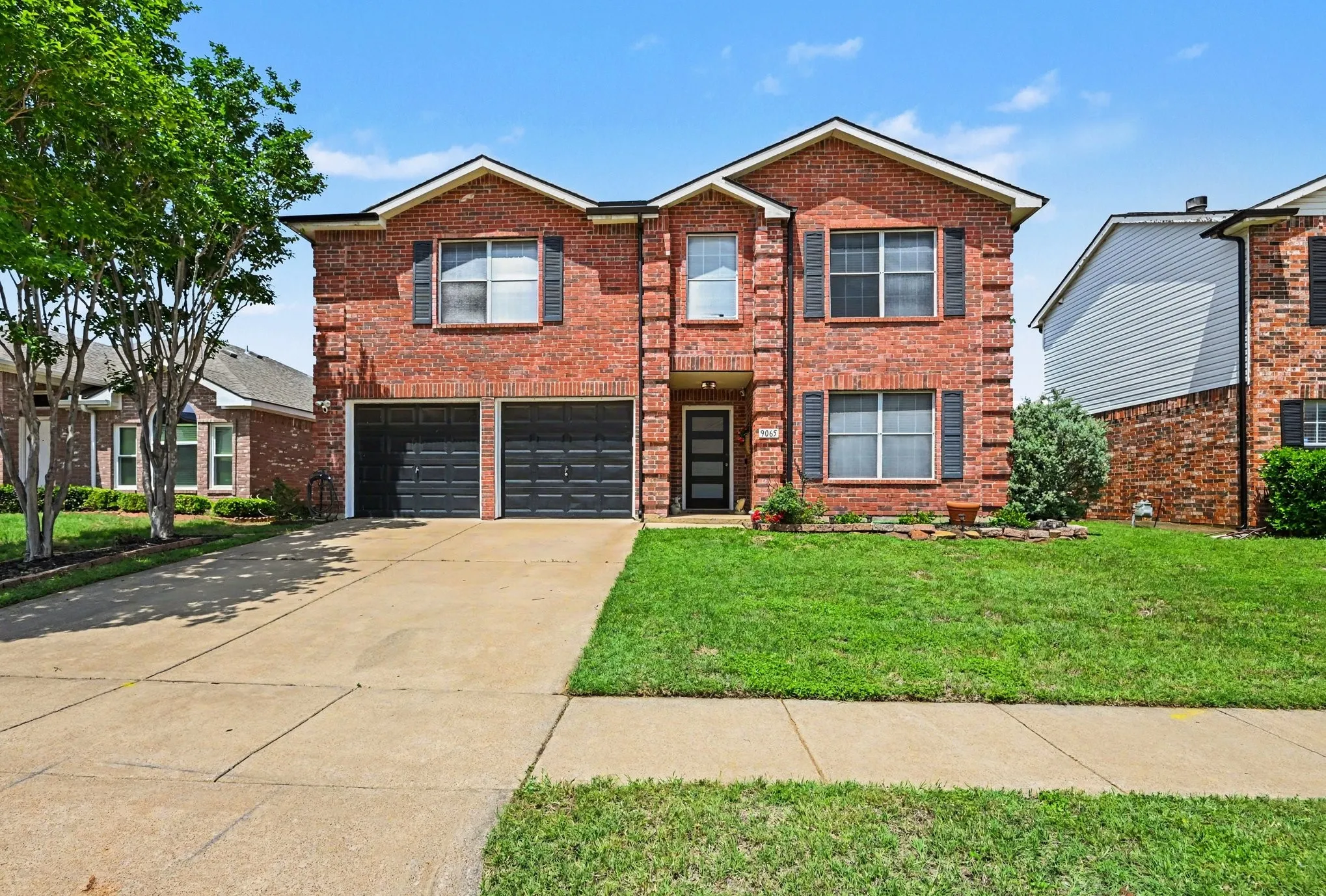 Striking two-story brick Colonial featuring a double-garage with dark panel doors, manicured front lawn with mature landscaping, concrete driveway and walkways, and multiple windows across the facade providing natural light throughout