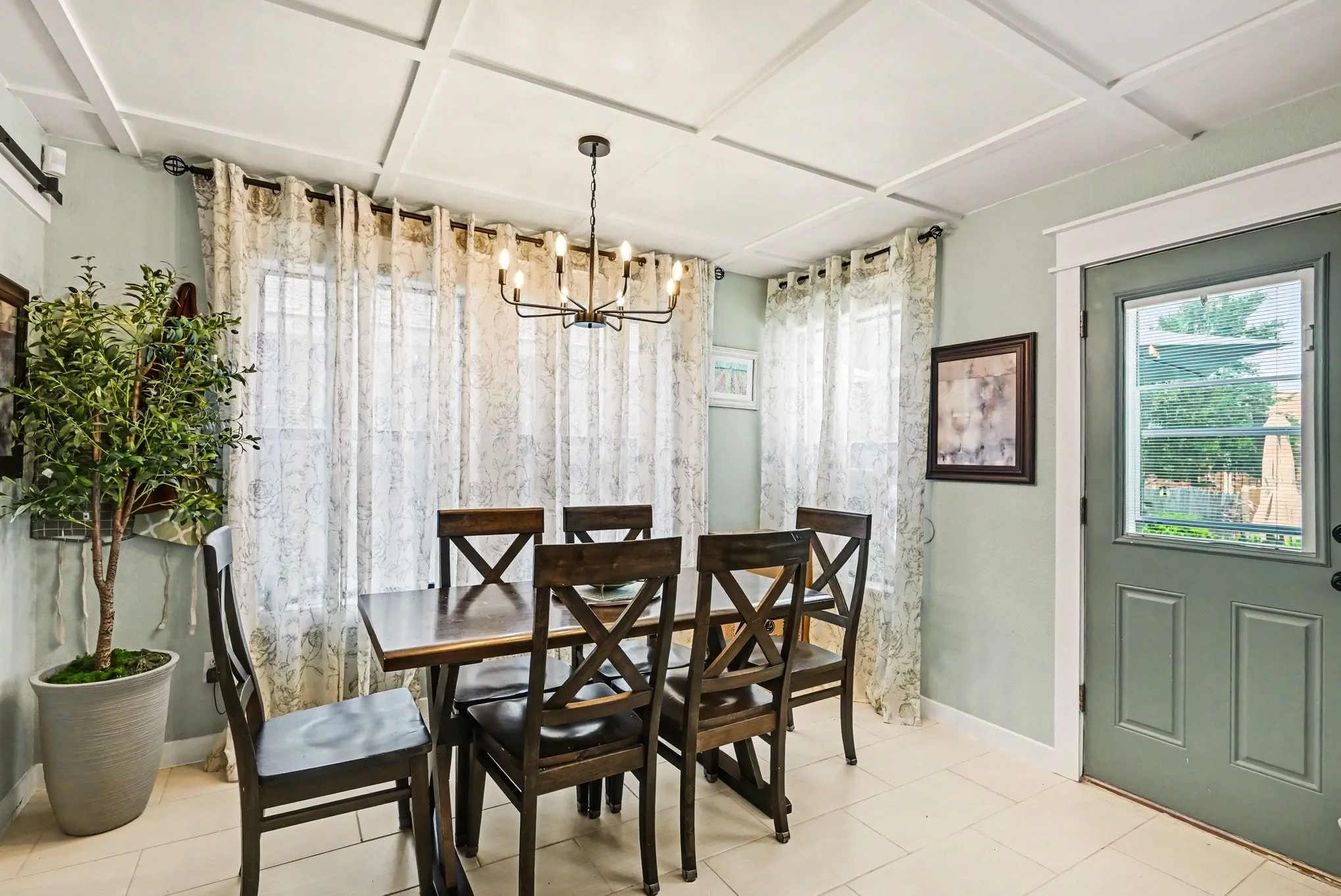 Elegant dining room featuring a coffered ceiling with pendant lighting, multiple windows with sheer curtains flooding the space with natural light, hardwood flooring, and a green six-panel entry door