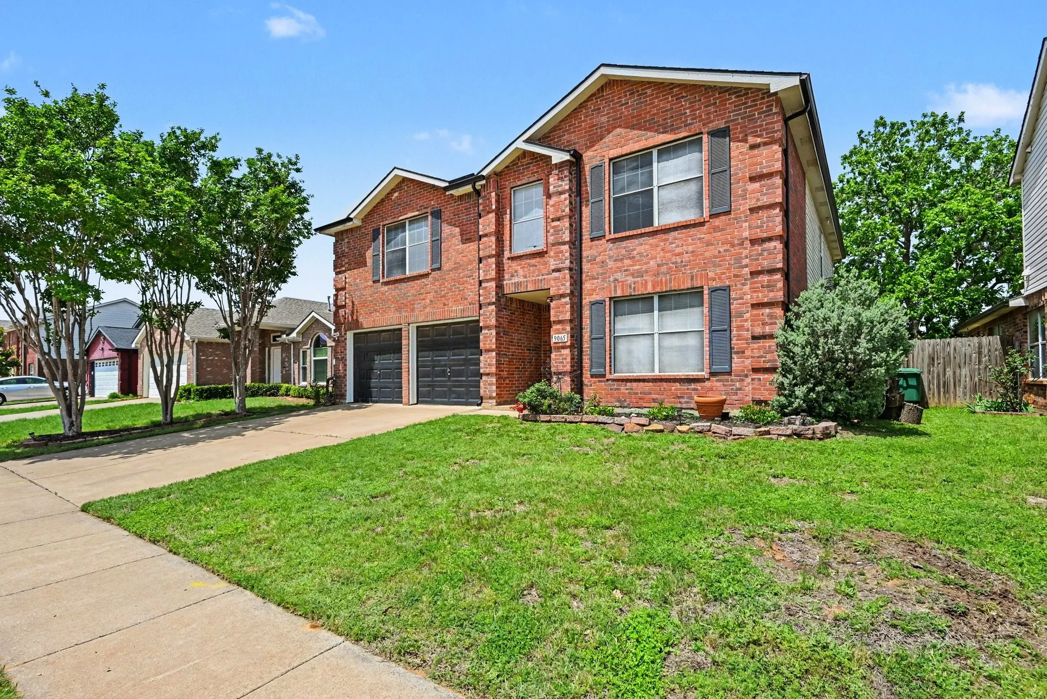 Brick exterior with dark shutters, two-car garage, concrete driveway, and established landscaping