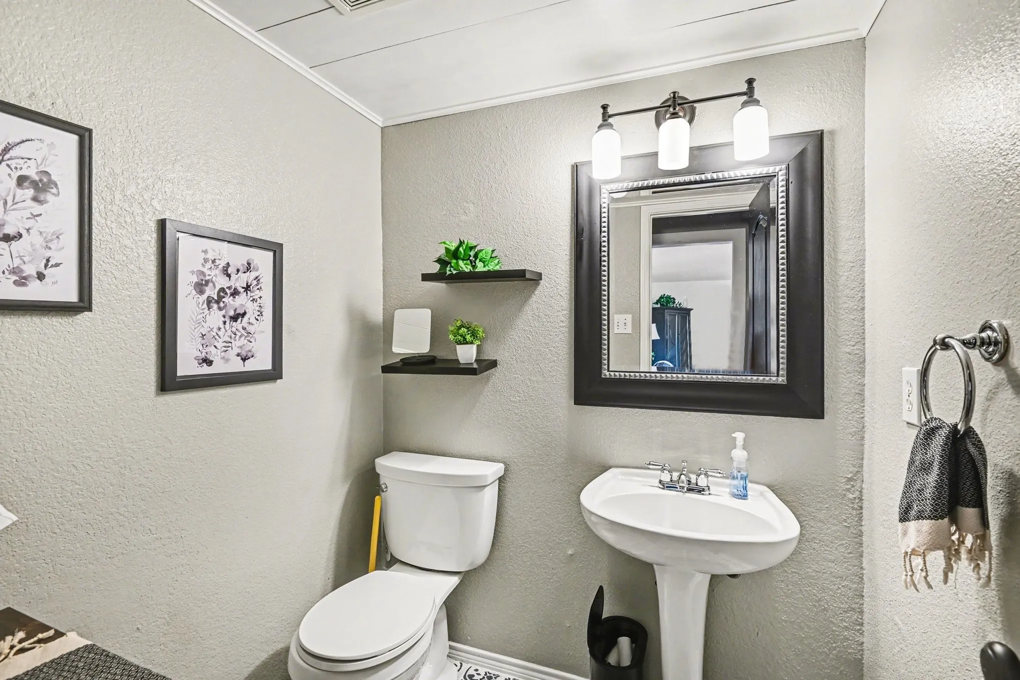 Pedestal sink, wall-mounted mirror with dark frame, three-light vanity fixture, white toilet, and floating wall shelves
