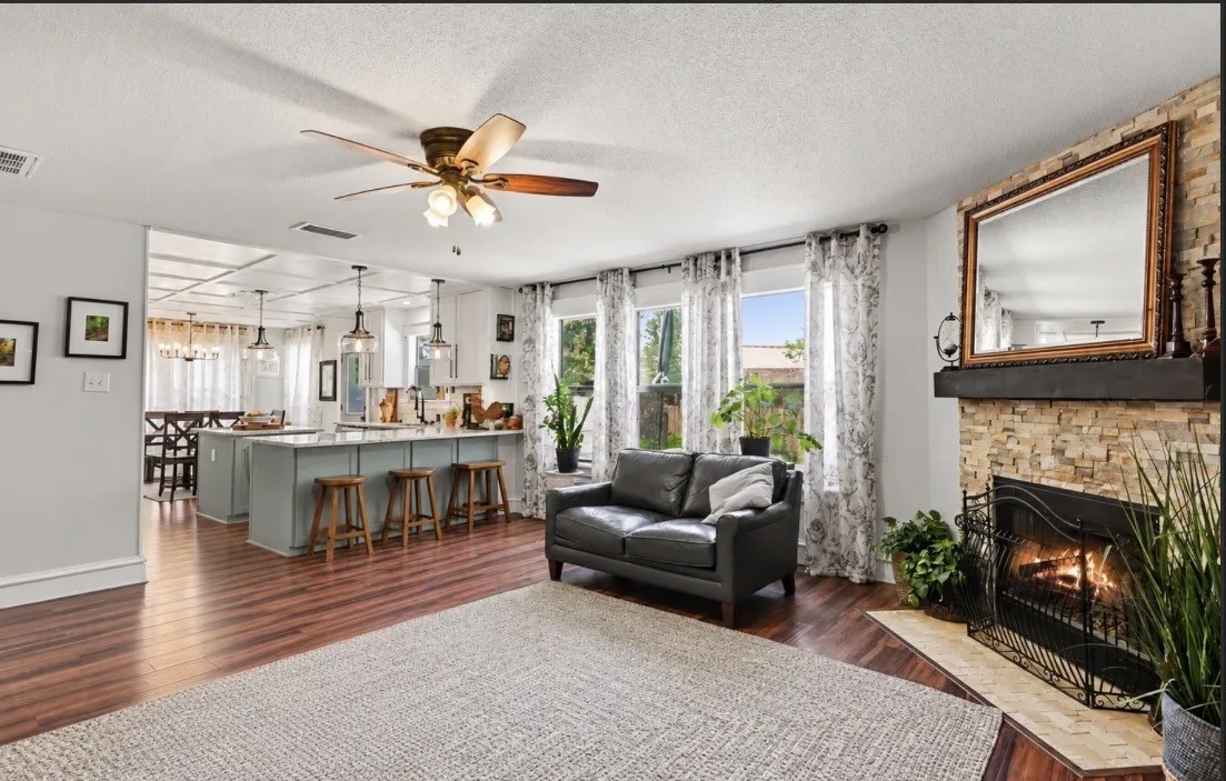Open concept living area featuring wood-finish flooring, a stacked stone fireplace, a ceiling fan with integrated lighting, and multiple windows