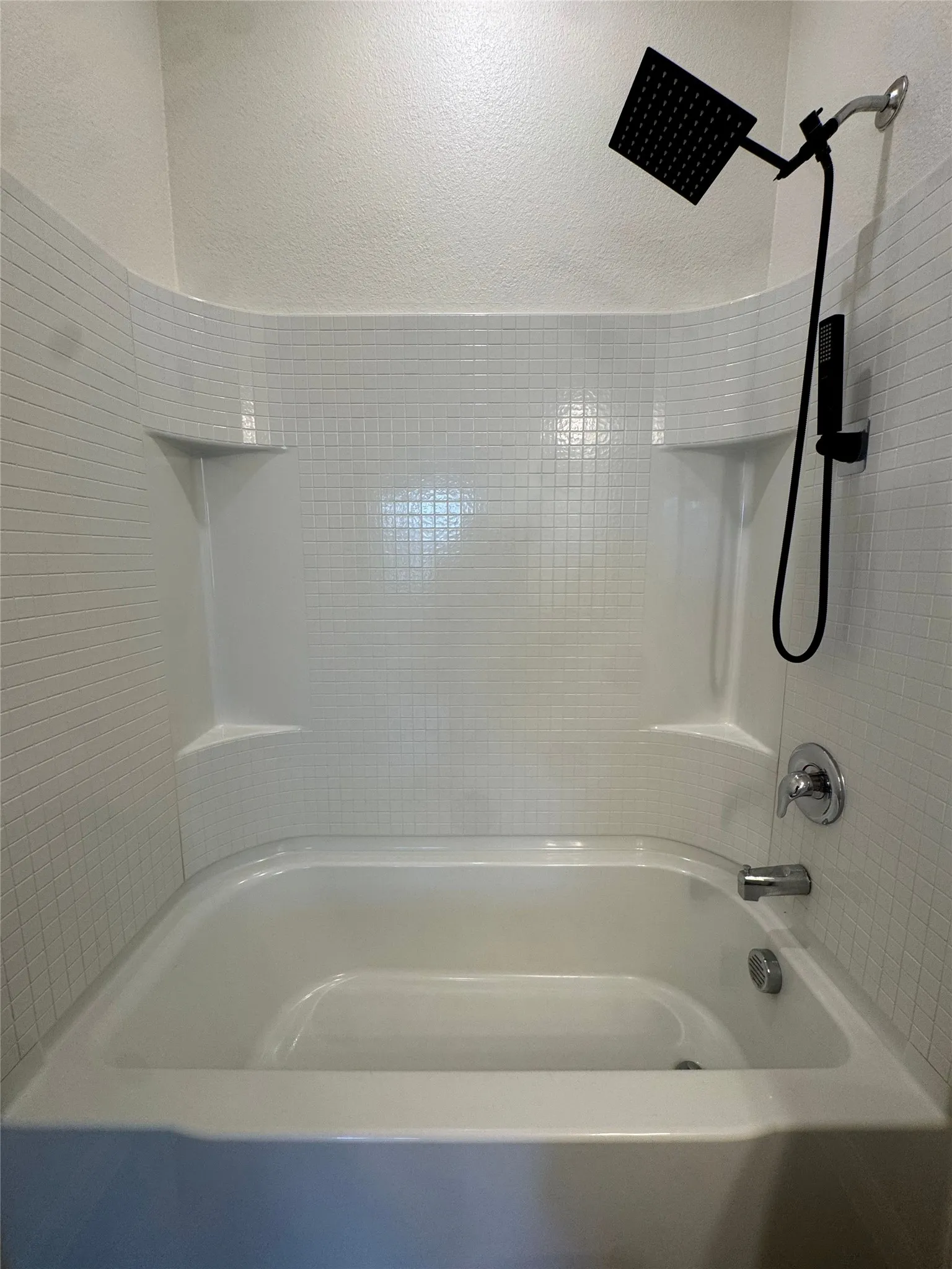 White bathtub with integrated shelving, white subway tile surround, and a black rain shower head