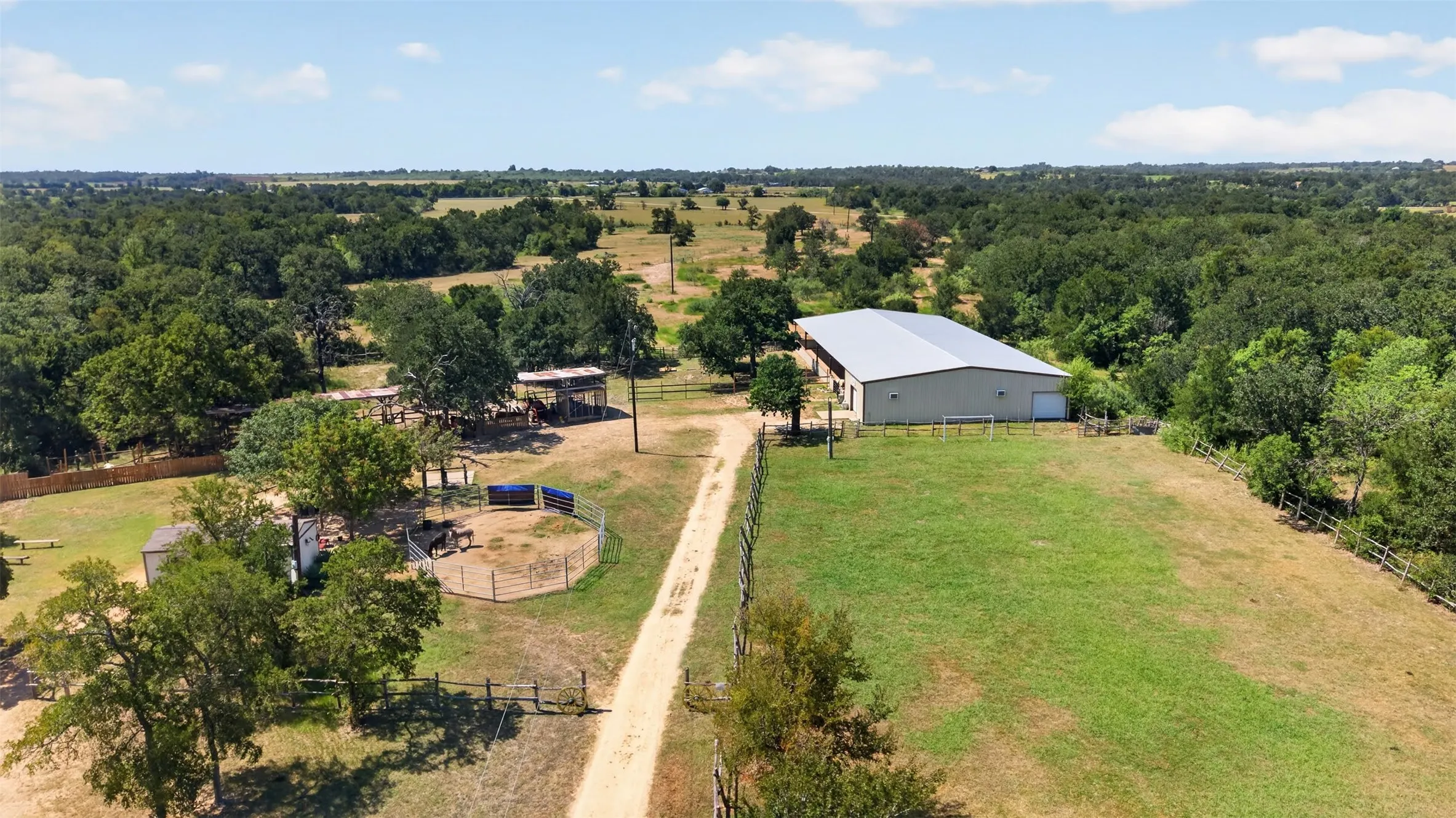 Sprawling rural property featuring a spacious metal-frame agricultural building with white exterior, expansive manicured pasture, mature tree-lined perimeter, and a long gravel driveway providing direct property access