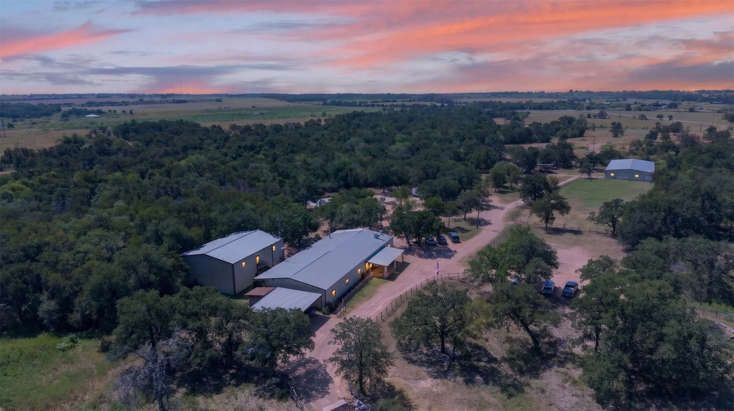 Expansive rural property featuring modern agricultural structures with metal roofing, situated on sprawling acreage surrounded by mature native trees and open pastureland