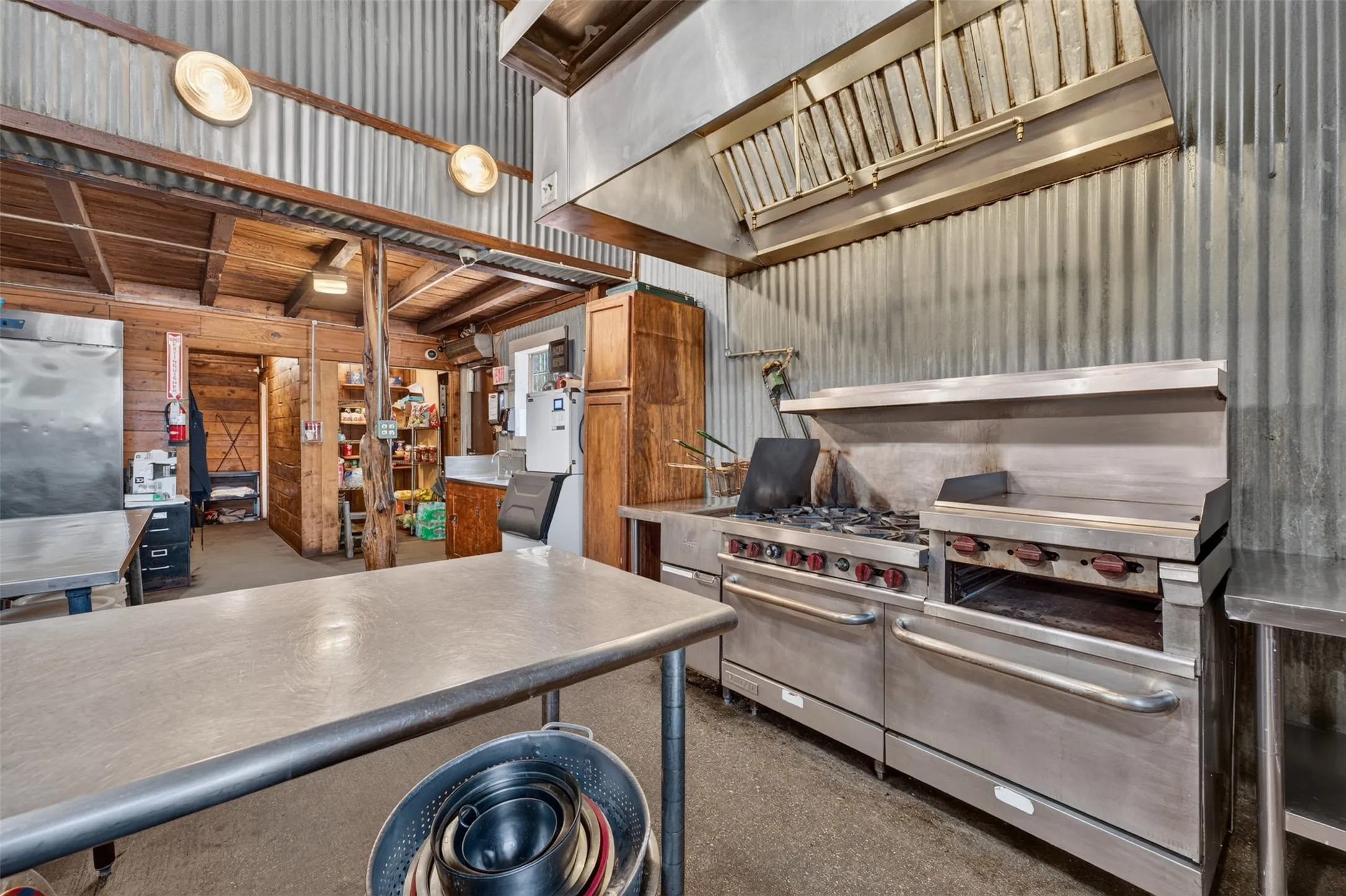 Professional kitchen featuring commercial-grade stainless steel range with multiple burners and integrated griddle, concrete countertops, wood cabinetry, and corrugated metal and wood-beam ceiling details