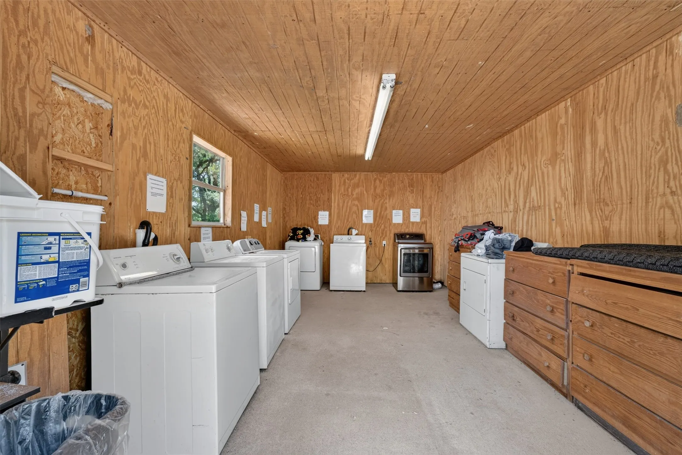Laundry facility featuring multiple commercial-grade washers and dryers, wood-paneled walls and ceiling, concrete flooring, and ample storage with wooden cabinetry and shelving throughout