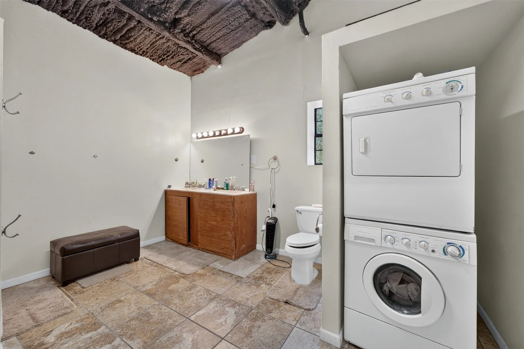 Tiled flooring, stacked laundry appliances, vanity with a rectangular mirror and light bar, and an exposed wood-finish ceiling