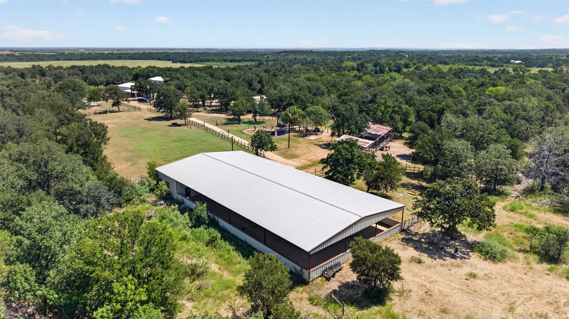 Modern agricultural or equestrian facility featuring a expansive white metal-roof structure with dark exterior framing, set on substantial acreage surrounded by mature trees and native vegetation