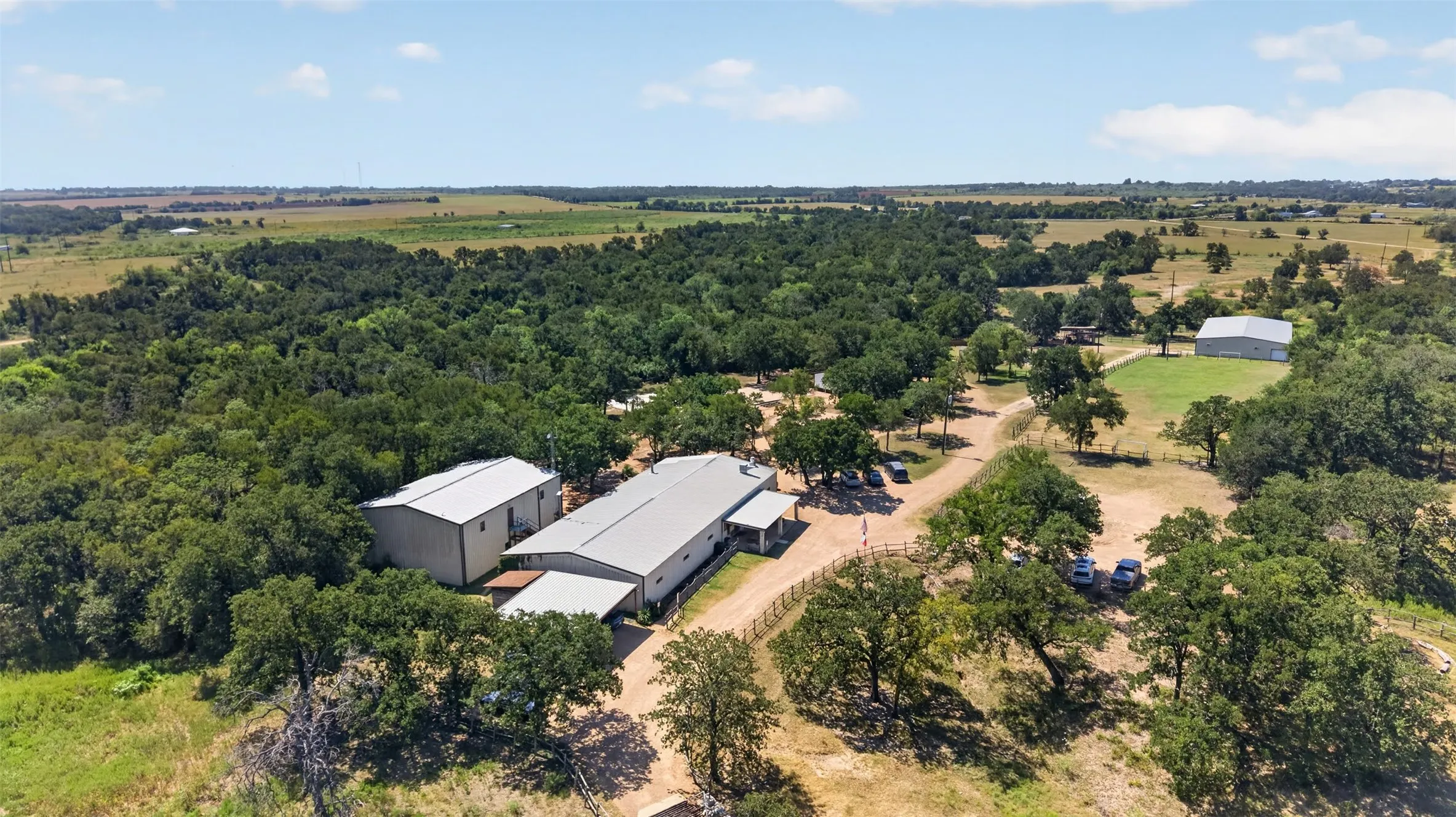 Expansive lot featuring mature trees, a gravel driveway, multiple metal-roofed structures, and open fields
