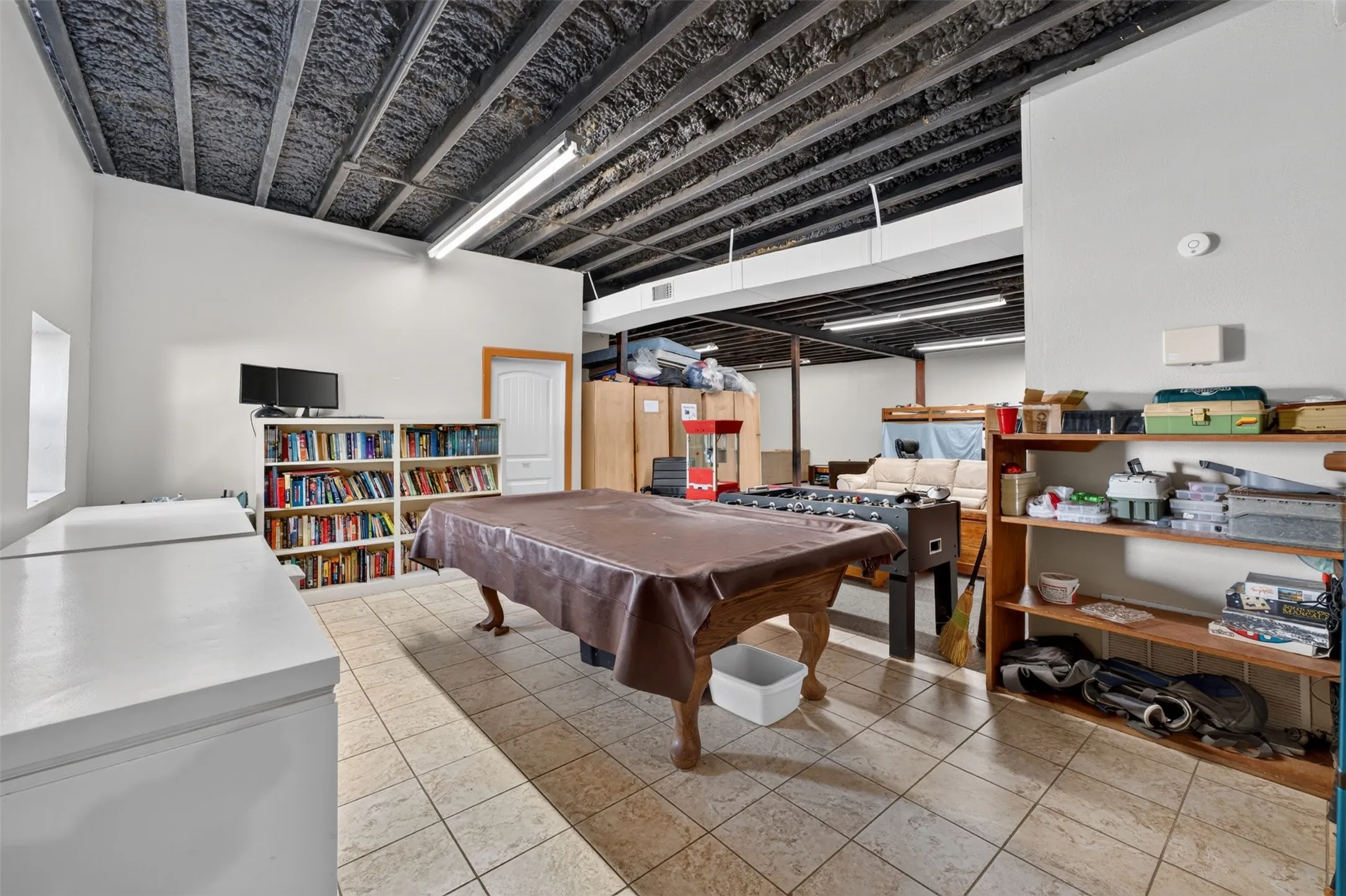 Versatile basement recreation space featuring exposed beam ceiling with industrial detailing, tile flooring, built-in wooden shelving and storage units, pool table, and ample room for multiple uses