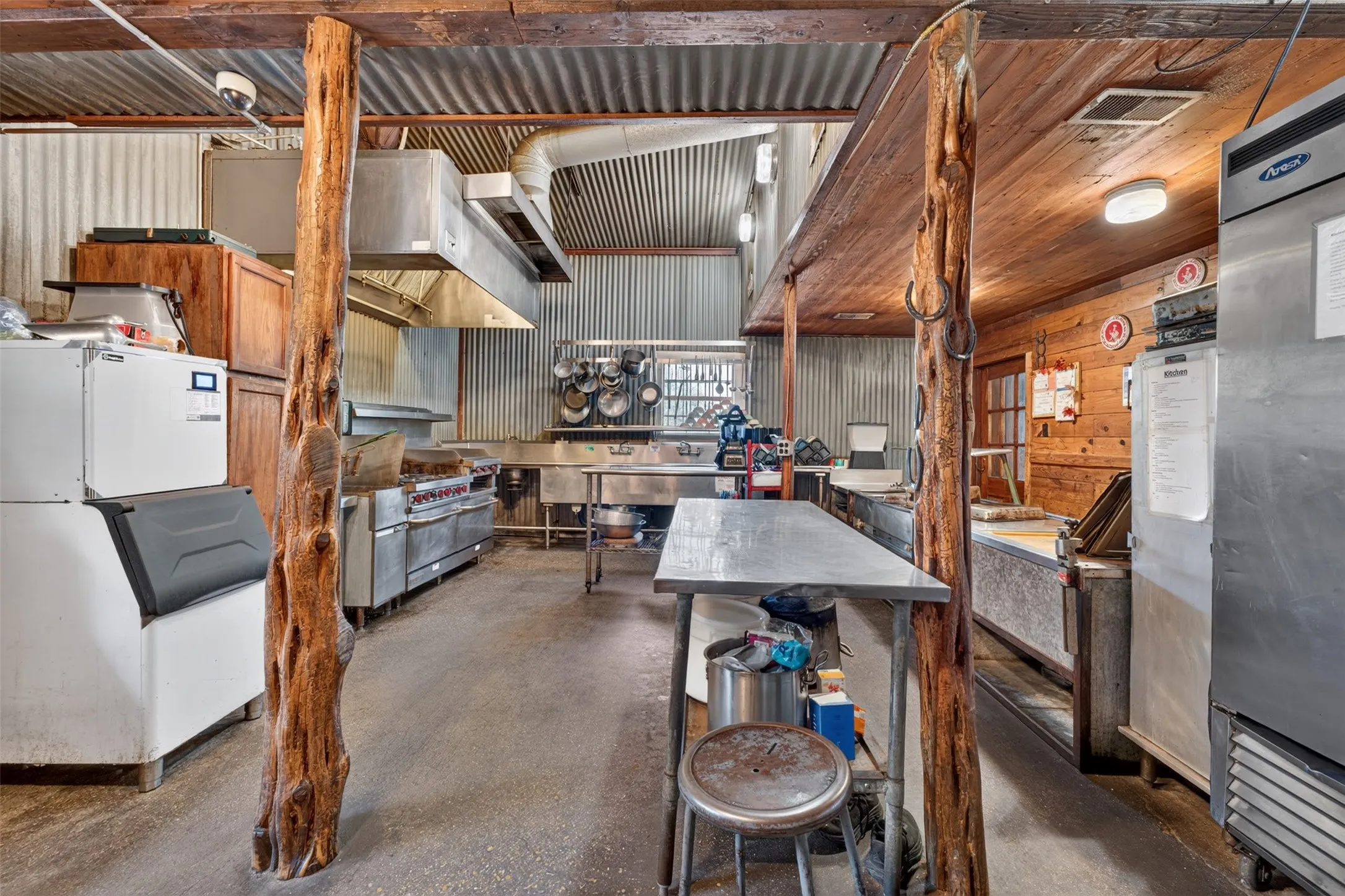 Commercial kitchen featuring corrugated metal walls, wood ceiling beams, stainless steel appliances, and concrete flooring