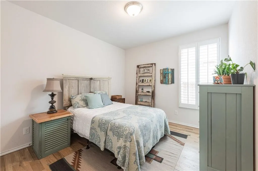 Bright secondary bedroom featuring wood-finish flooring, sage green built-in cabinetry with potted plants, paired nightstands with task lighting, and natural light from multi-pane windows with plantation shutters