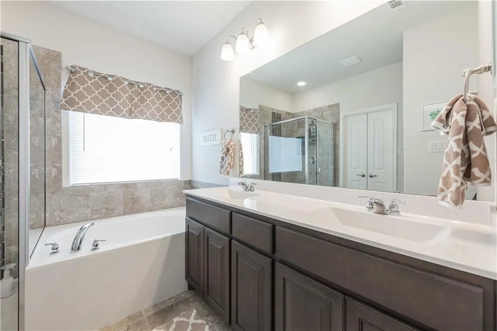 Spa-inspired master bathroom featuring a dual-sink vanity with dark cabinetry and white quartz countertops, large framed mirror, soaking tub with tile surround, walk-in shower with glass enclosure, and tile flooring throughout