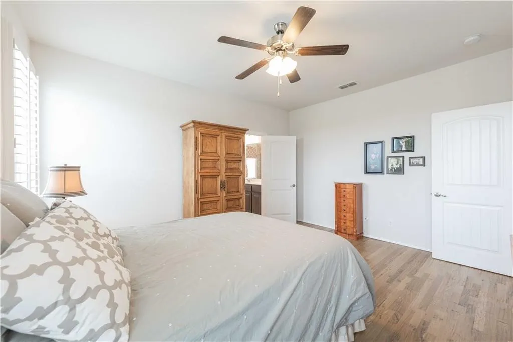Spacious primary bedroom showcasing wood-finish flooring, ceiling fan with integrated lighting, and abundant natural light from shuttered windows