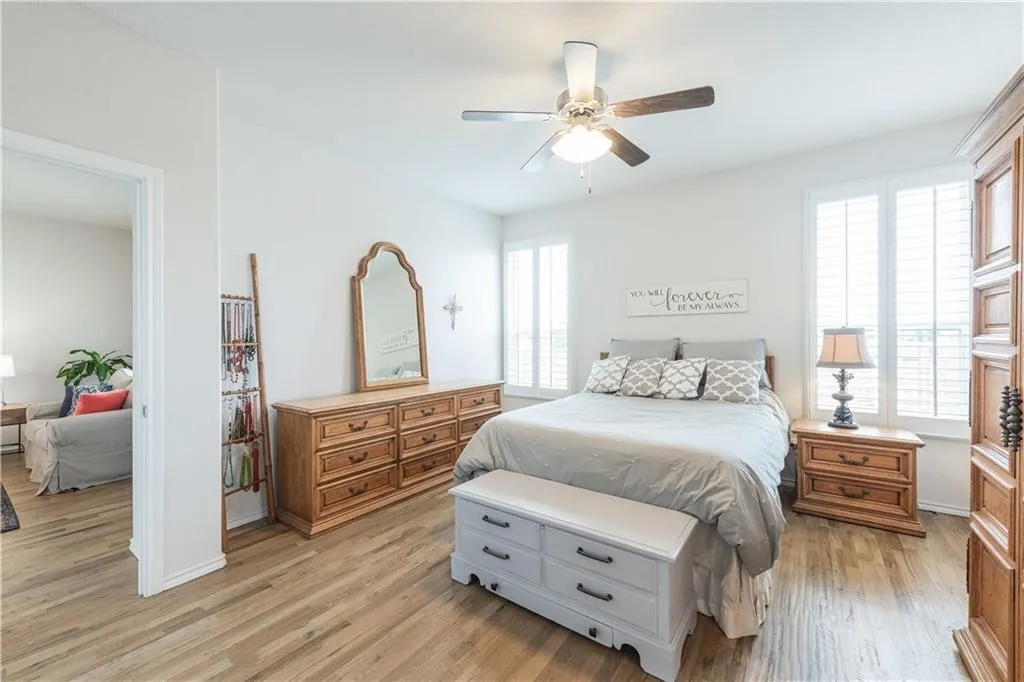 Spacious primary bedroom featuring hardwood flooring, ceiling fan with integrated lighting, wooden dresser with mirror, nightstands with lamps, and abundant natural light from multiple windows and glass doors overlooking the outdoor space