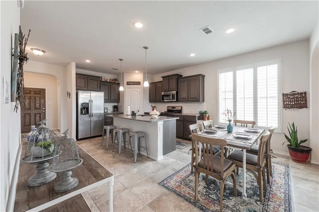 Open-concept kitchen and dining area featuring dark cabinetry, stainless steel appliances, a large central island with seating, tile flooring, recessed lighting, and abundant natural light from French doors opening to the exterior