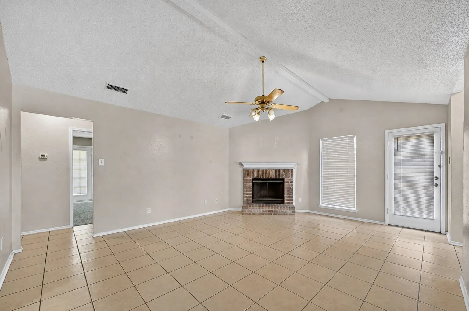 Vaulted ceiling with exposed beams, brick fireplace with mantel, tiled flooring, ceiling fan with integrated lighting, and a glass-paneled door