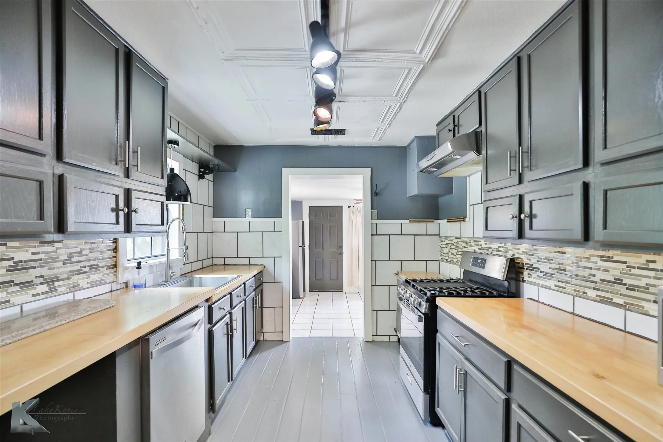 Galley-style kitchen featuring gray cabinetry with glass-front upper cabinets, white subway tile backsplash with contrasting grout lines, wood-finish countertops, stainless steel gas range, and white tile flooring throughout