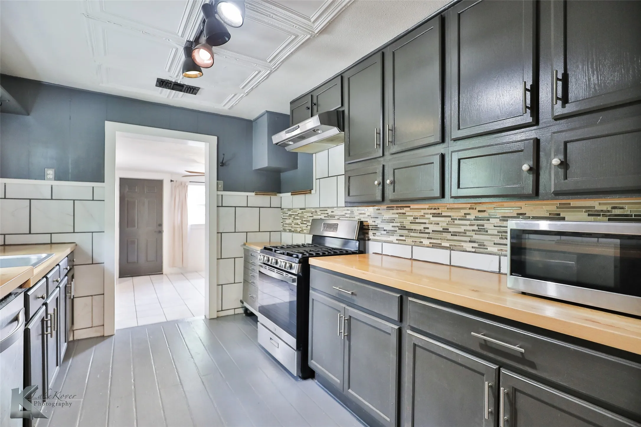 Kitchen featuring painted wood flooring, dark cabinetry, light wood-finish countertops, subway tile wainscoting, and a mosaic tile backsplash