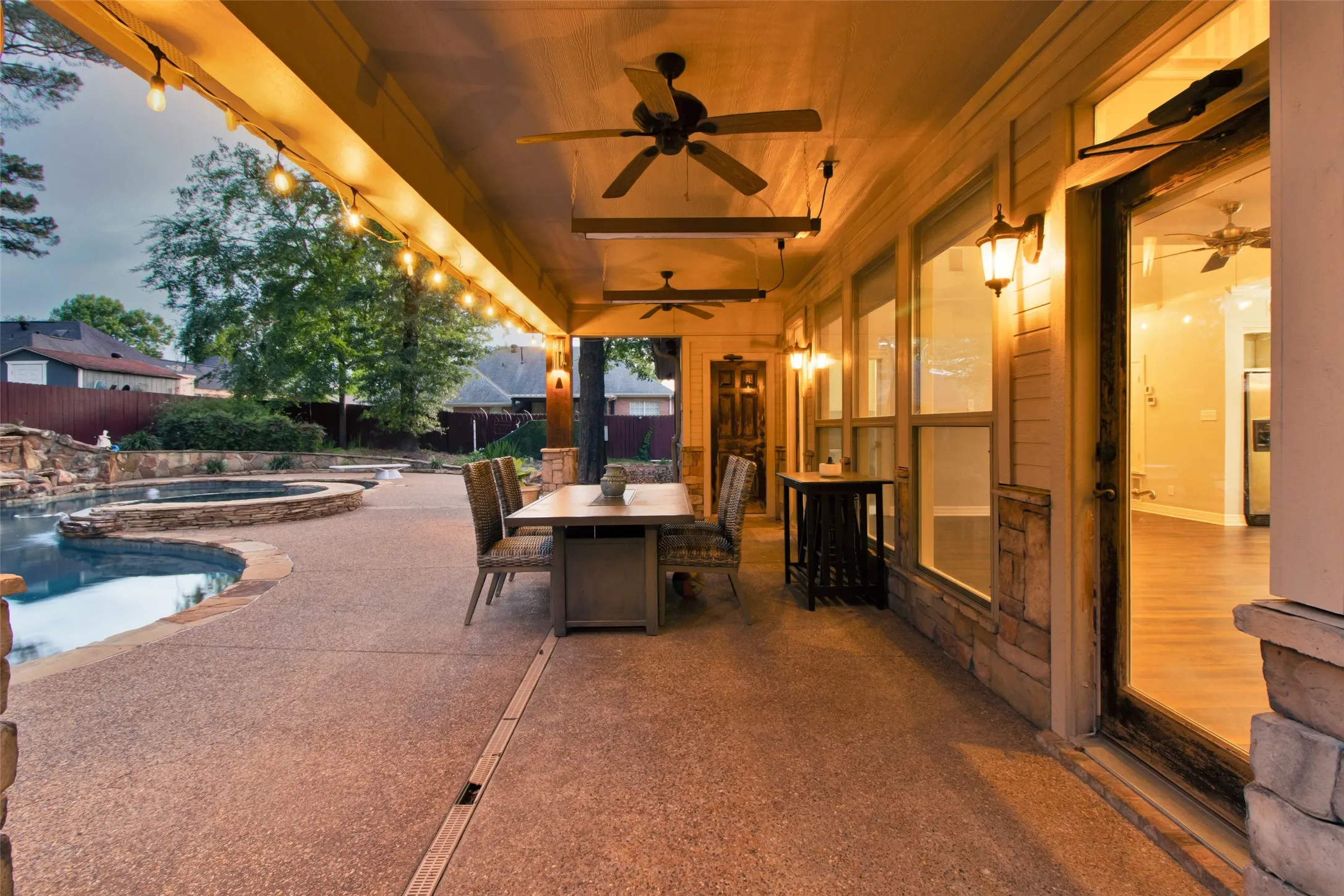 Covered patio with exposed aggregate concrete flooring, featuring multiple ceiling fans, string lighting, and direct access to a swimming pool with a stone-accented spa