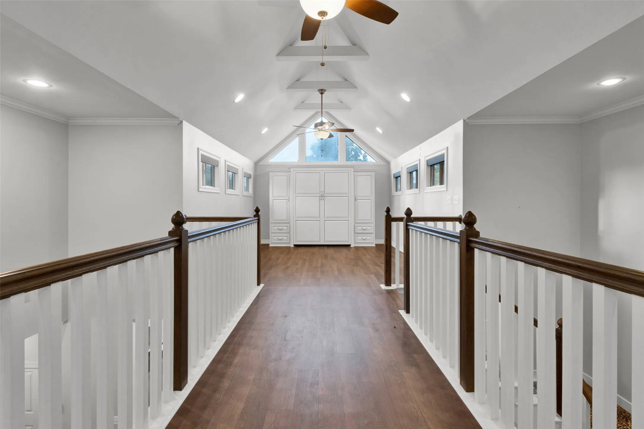 Spacious loft area featuring wood-finish flooring, a vaulted ceiling with exposed beams, multiple ceiling fans with integrated lighting, recessed lighting, and a built-in white cabinetry unit with a triangular window above