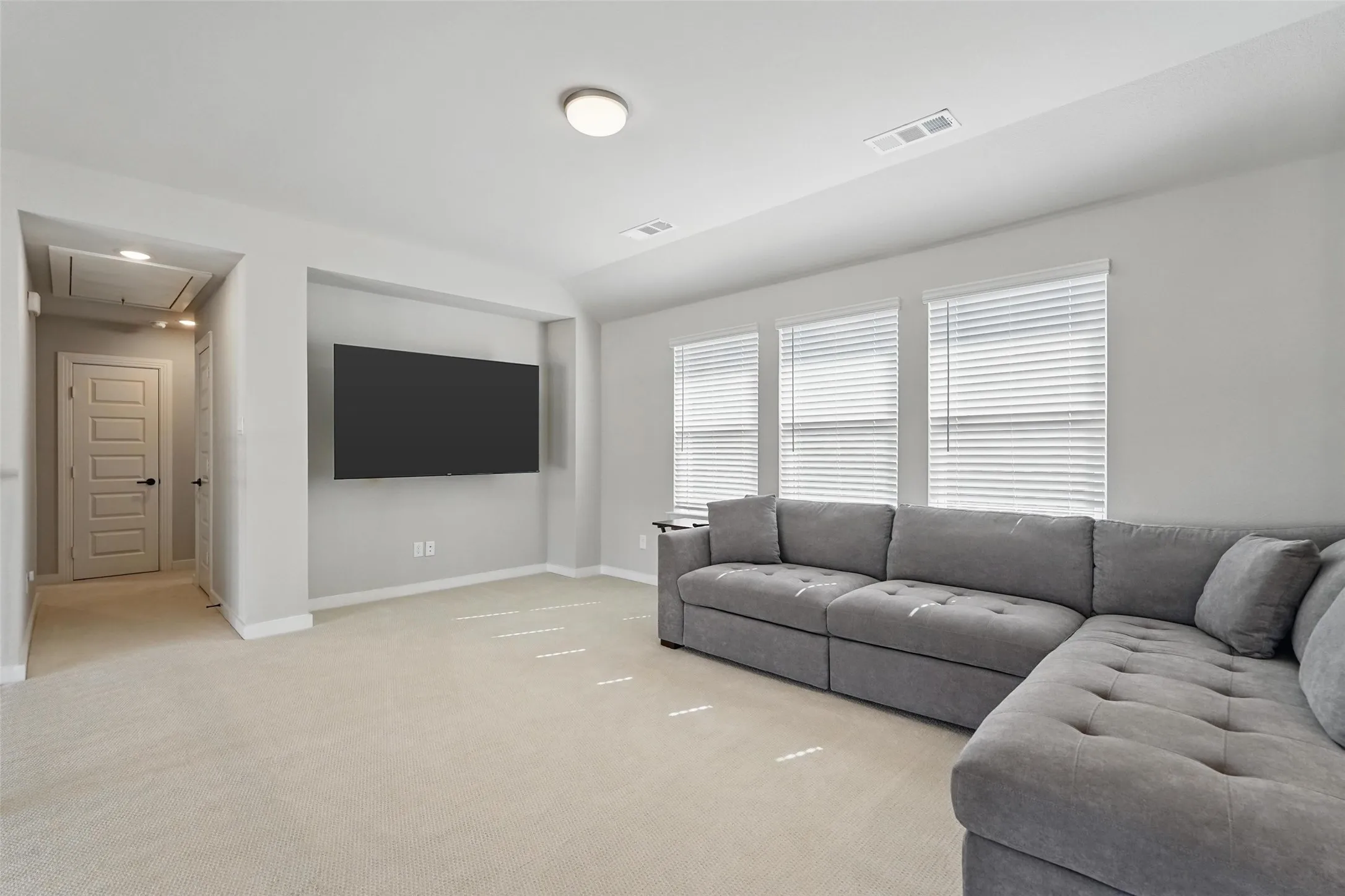 Recessed wall feature for television, three windows with blinds, light-toned carpet flooring, overhead flush-mount lighting, and a paneled interior door
