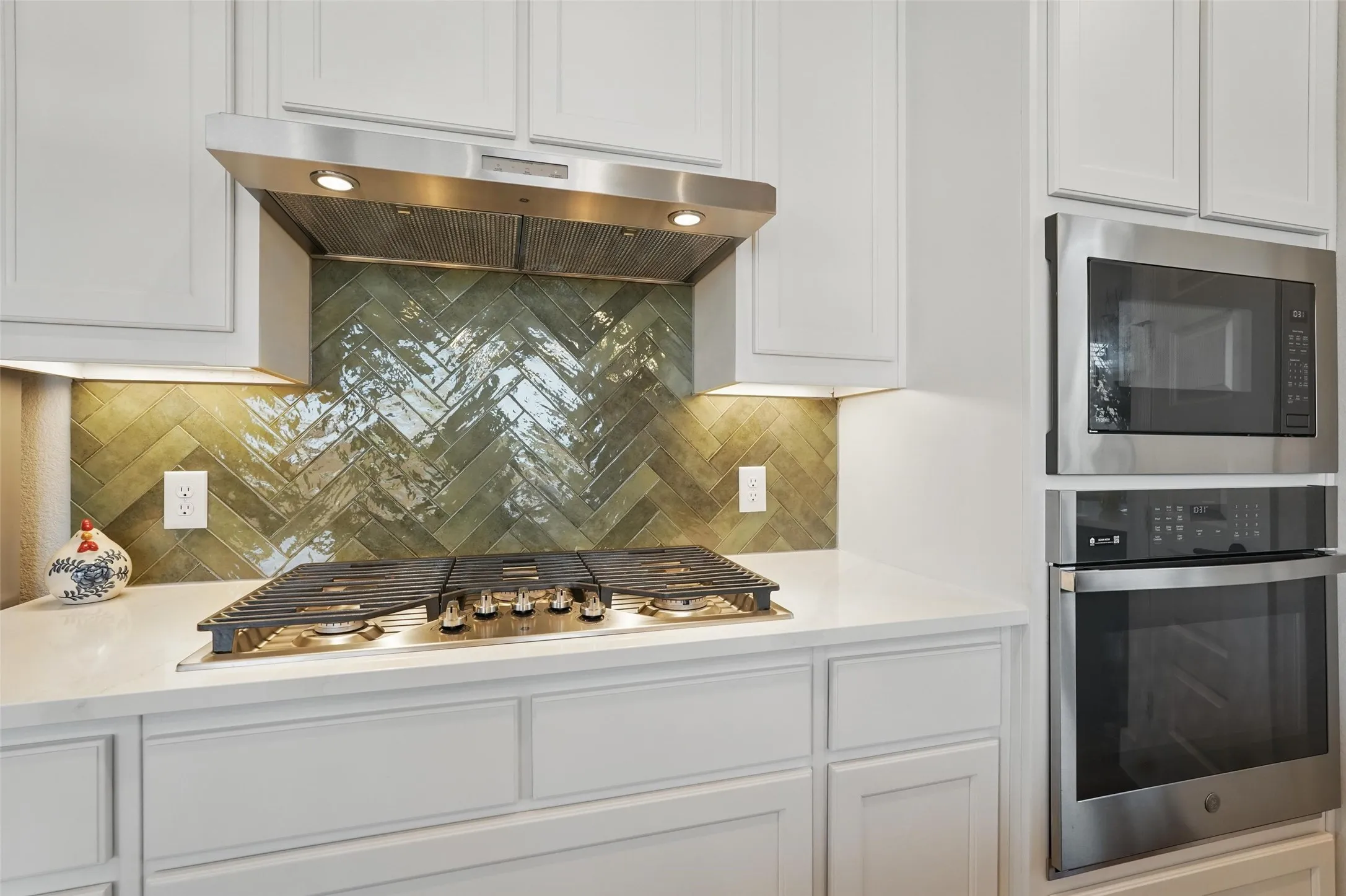 Built-in gas cooktop, stainless steel range hood, herringbone tile backsplash, white shaker-style cabinetry, and built-in stainless steel microwave and oven