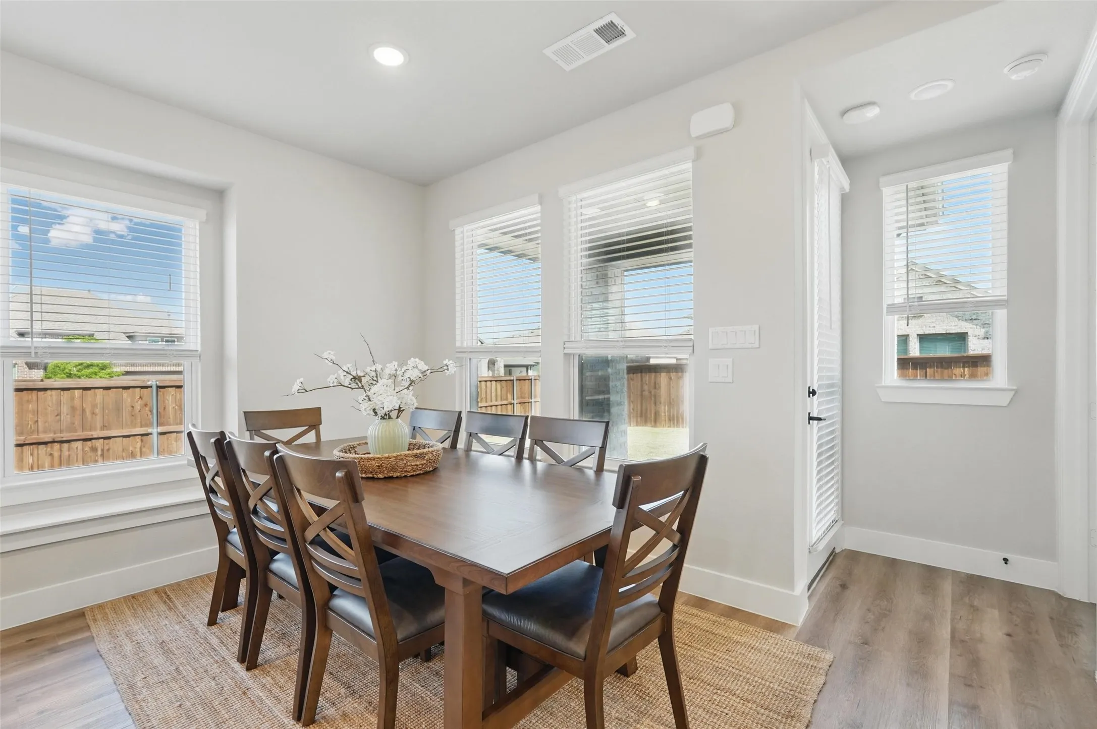 Bright dining space flooded with natural light through multiple windows and glass door, featuring a wood-finish dining table with seating for four, wide-plank wood-finish flooring, neutral wall finishes, and recessed ceiling lighting