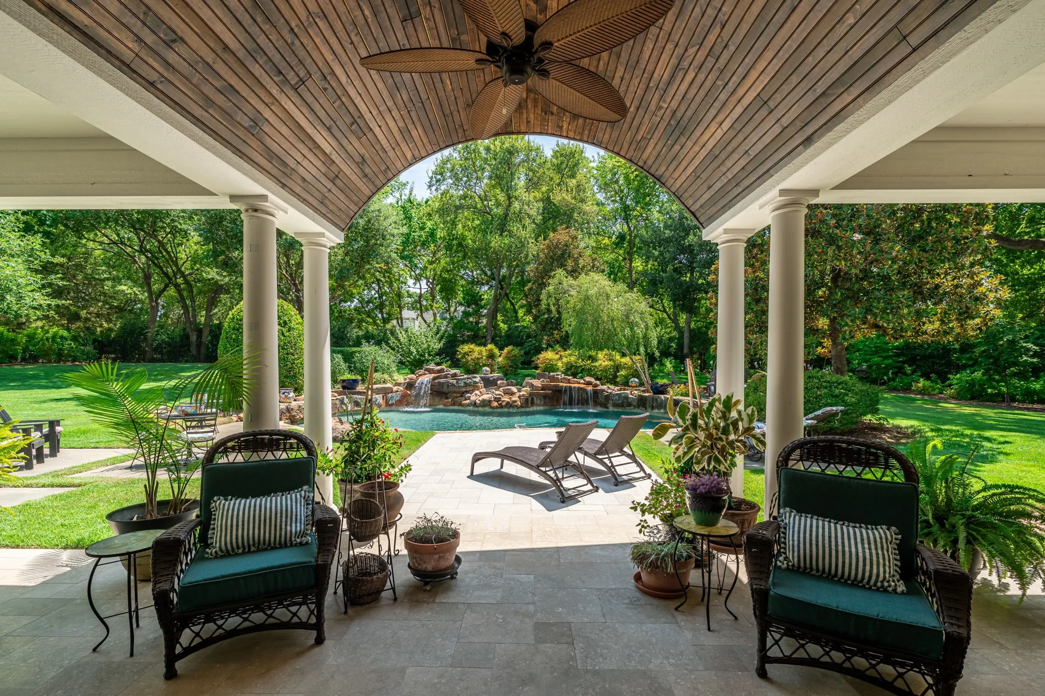 Covered loggia with wood-finish ceiling and ceiling fan opens to resort-style backyard featuring in-ground swimming pool, mature landscaping, expansive manicured lawn, and lounge seating areas