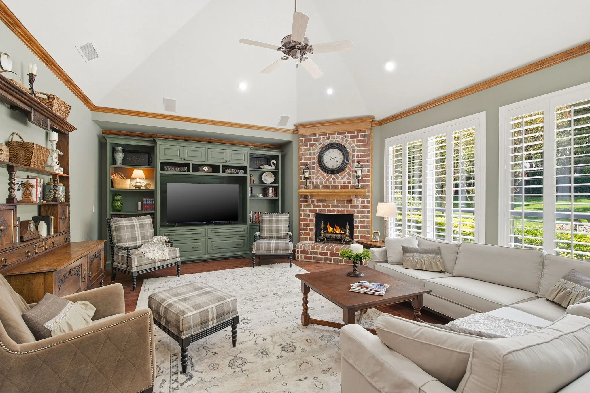 Vaulted ceiling with a ceiling fan, built-in entertainment cabinetry, brick fireplace with a wood mantel, and multiple windows with plantation shutters