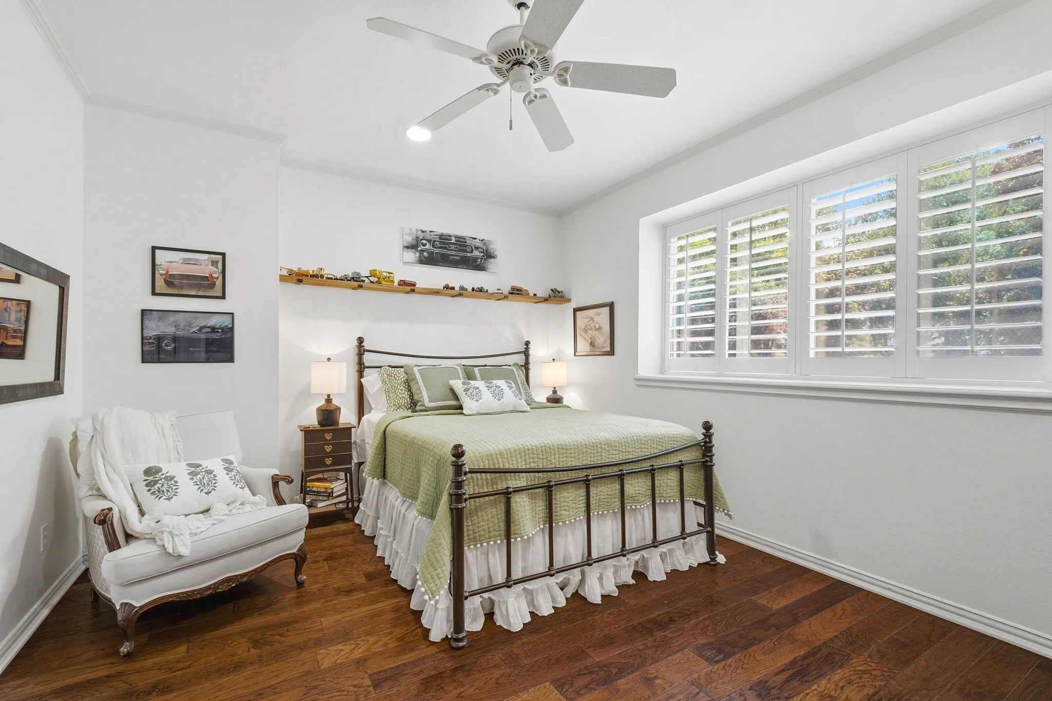 Bright secondary bedroom showcasing wood-finish flooring, a metal frame bed, plantation shutter window treatments, and a ceiling fan with integrated lighting