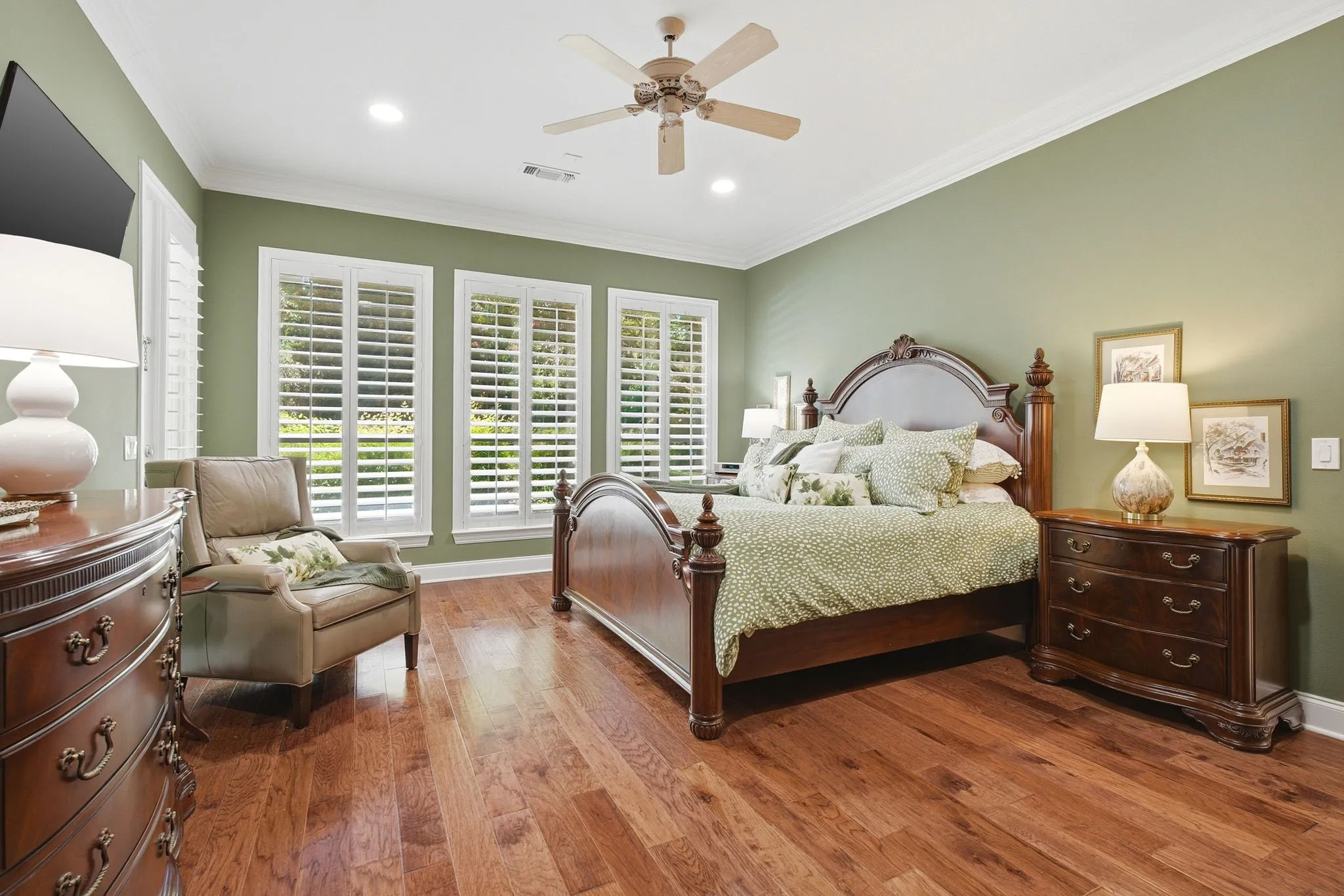 Primary bedroom suite featuring hardwood flooring, sage green walls, and triple plantation shutter-style closet doors with natural light