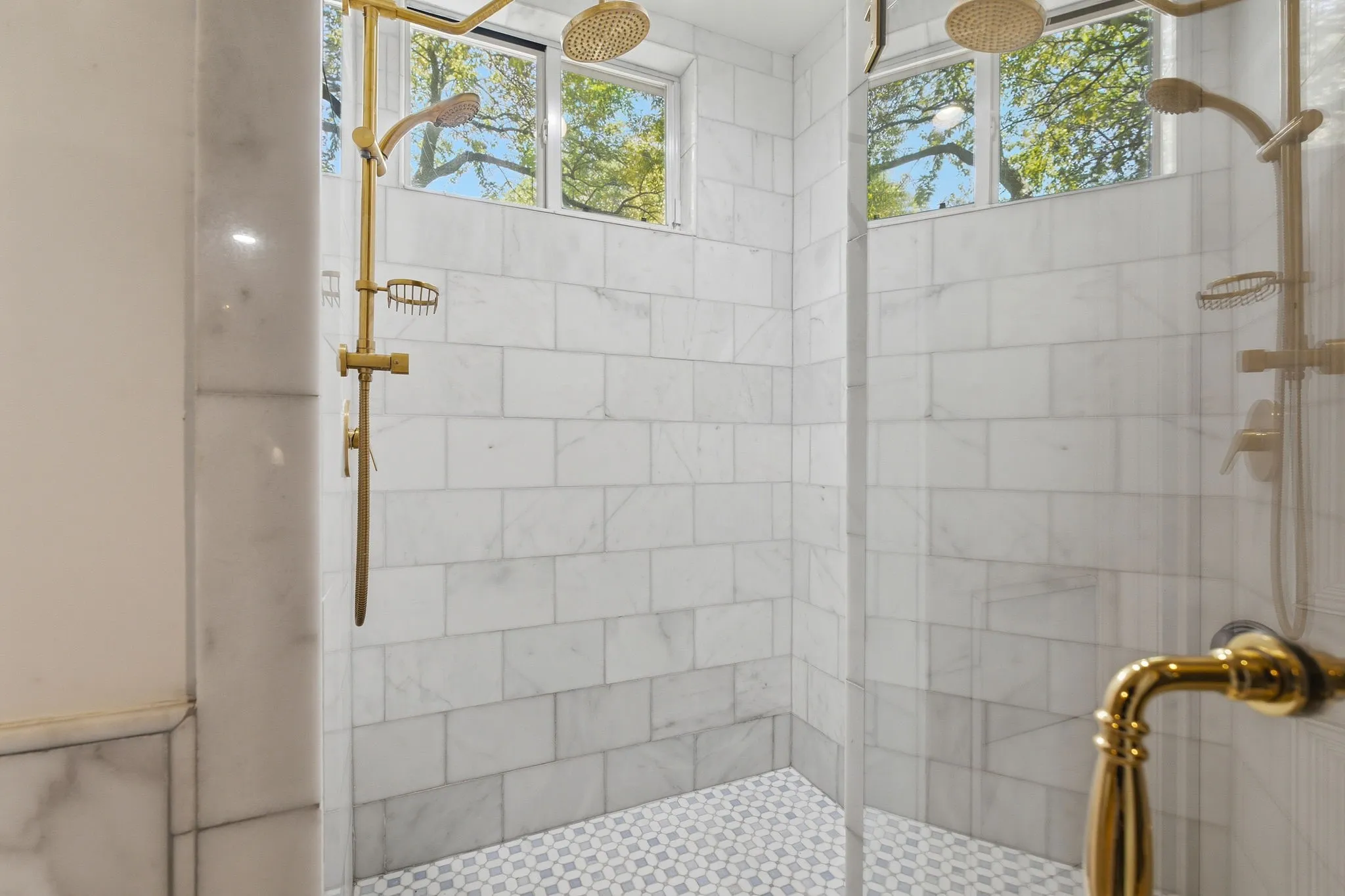 Spa-inspired primary bathroom featuring a walk-in shower with white subway tile walls, polished hexagonal mosaic floor, dual rainfall showerheads with brass fixtures, and transom windows providing natural light and ventilation