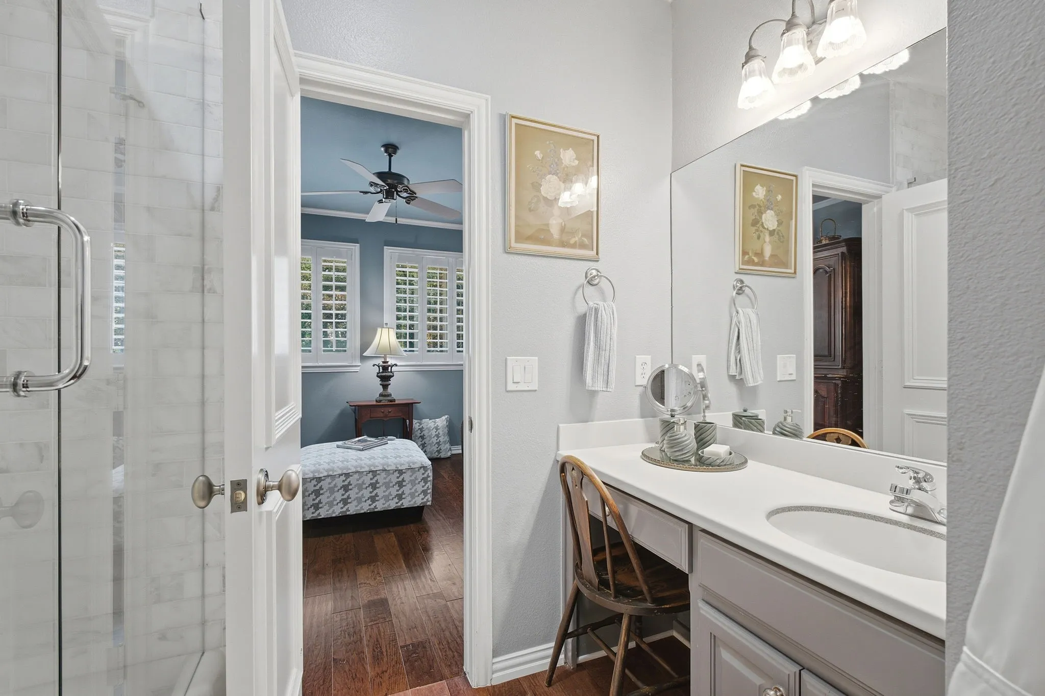 Spa-inspired ensuite bathroom featuring a frameless glass-enclosed shower with marble subway tile surround, dual vanity with white cabinetry and rectangular vessel sinks, polished nickel fixtures, LED vanity lighting, and access to the adjoining bedroom suite with hardwood flooring and ceiling fan