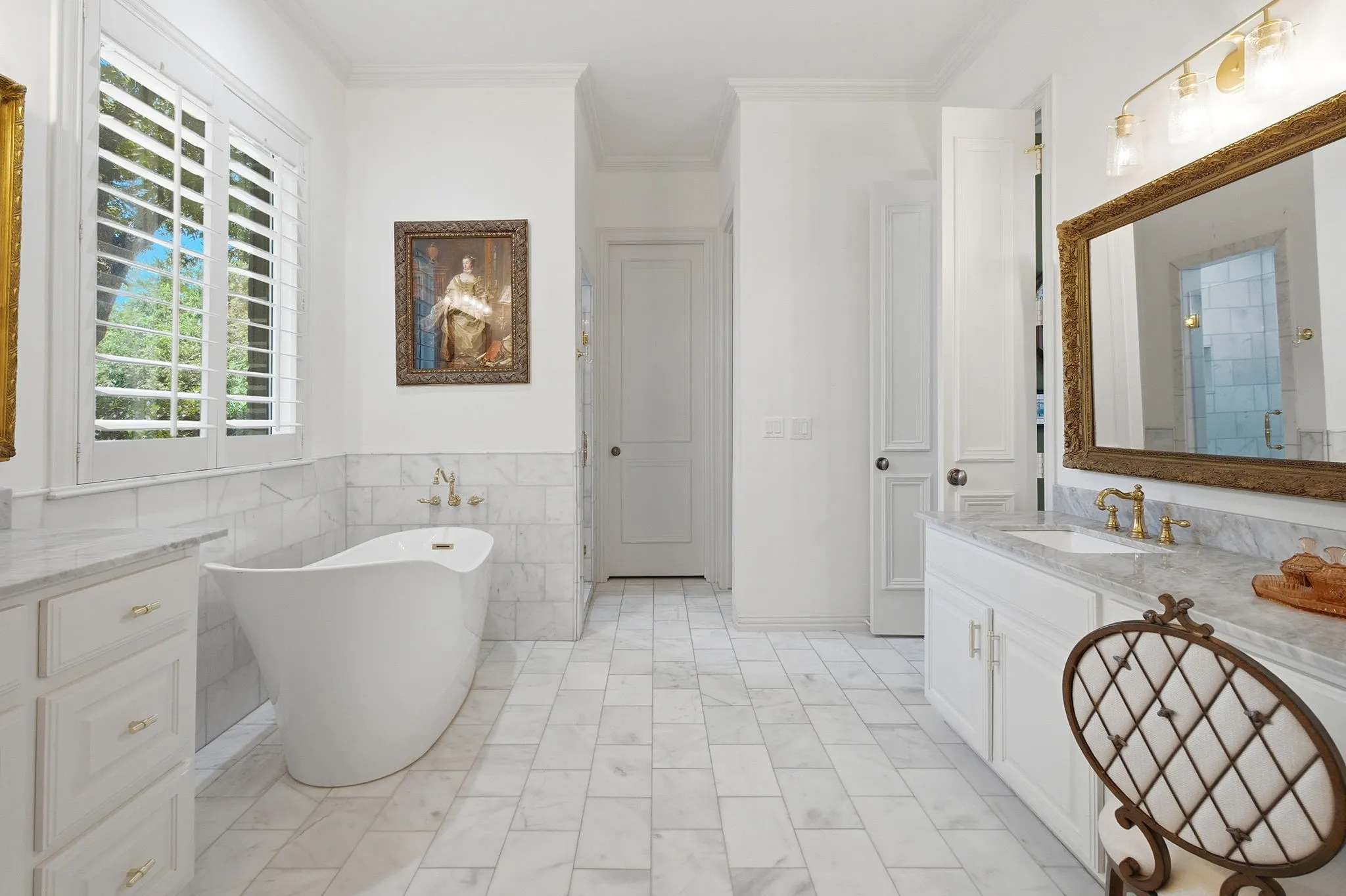 Spa-inspired primary bathroom featuring a freestanding soaking tub, dual vanities with marble-finish countertops and brass fixtures, large framed mirror, geometric tile flooring, plantation shutters, and abundant natural light from corner windows