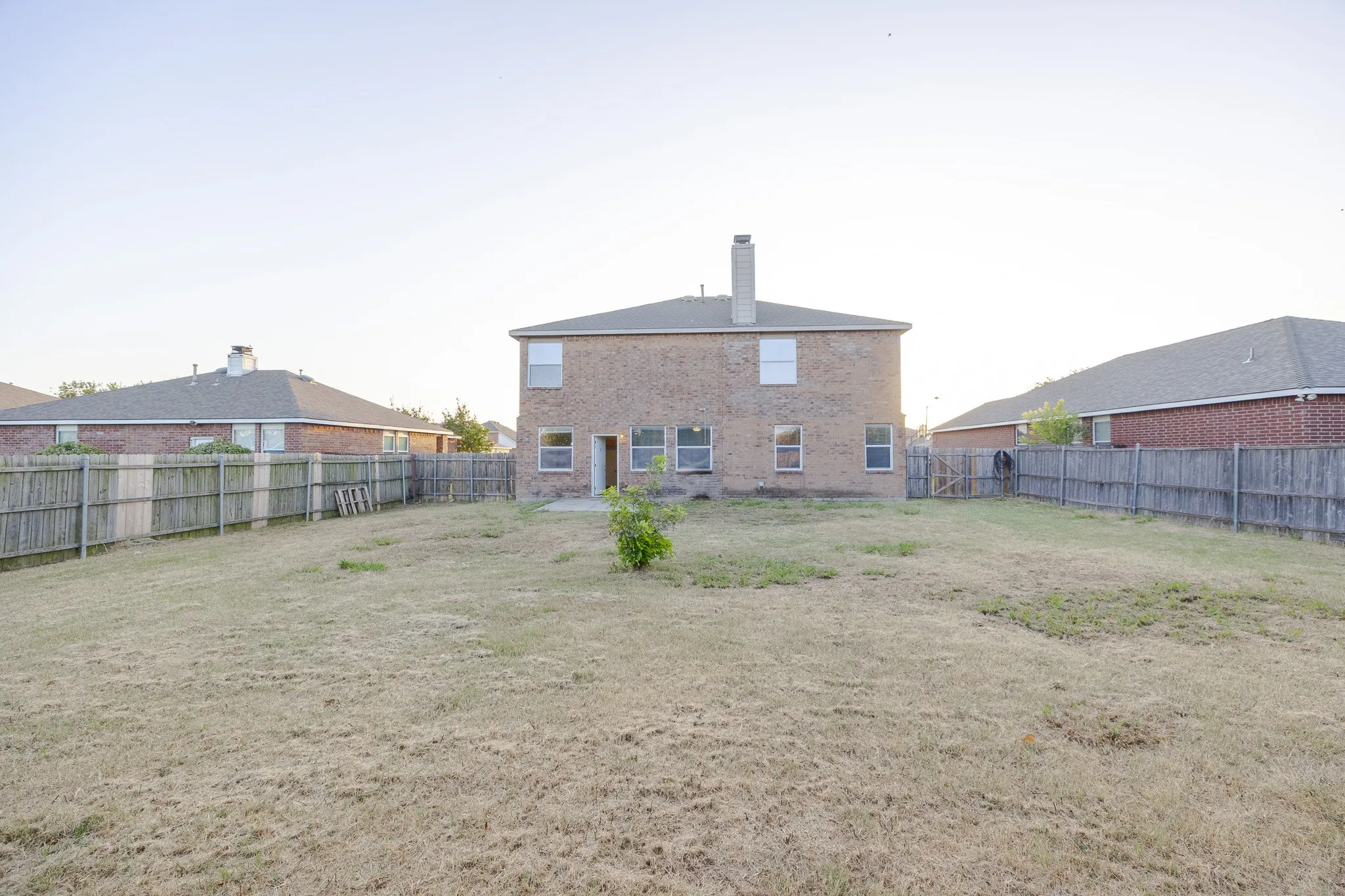 Spacious backyard with expansive lawn and fenced perimeter, anchored by a two-story brick residence featuring a hip roof, central chimney, and multiple window openings