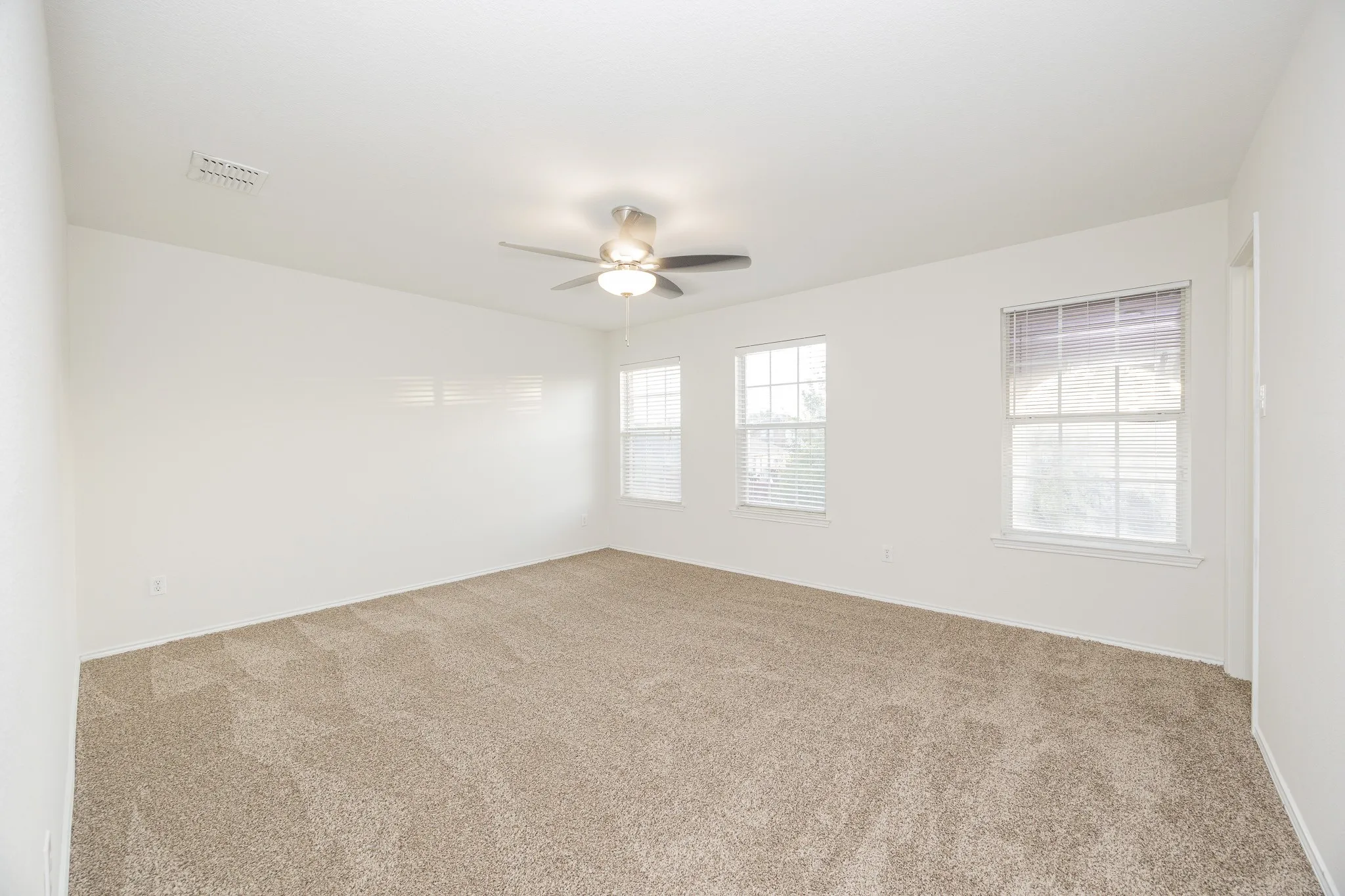 Spacious room featuring neutral carpet flooring, three windows with blinds, and a ceiling fan with integrated lighting
