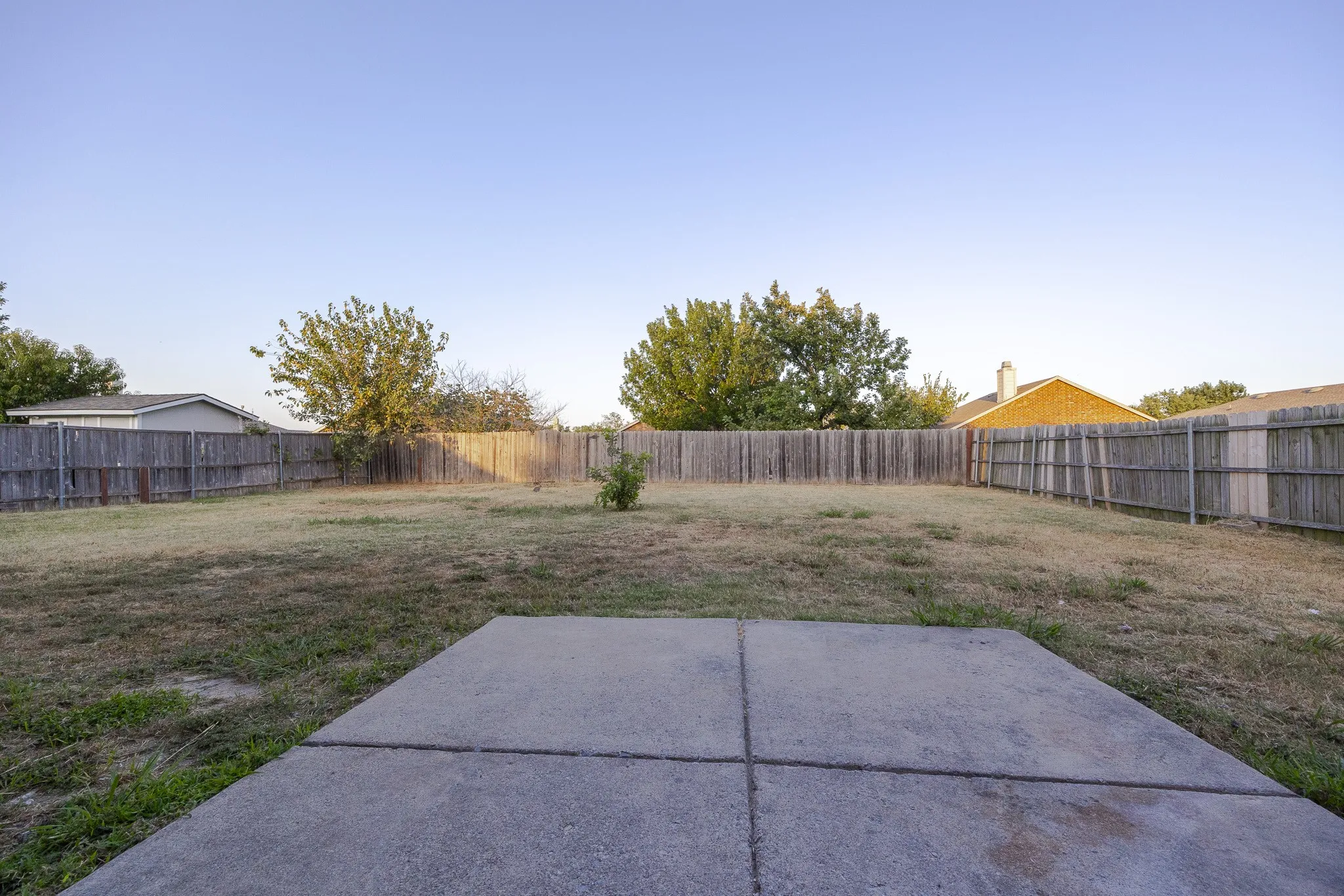 Expansive backyard featuring a concrete patio pad, mature shade trees, and wooden perimeter fencing
