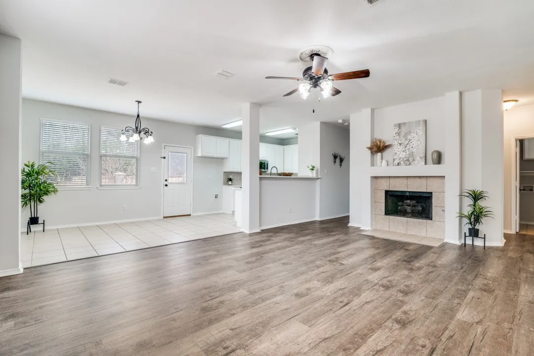 Wood-finish flooring, tiled fireplace with mantel, ceiling fan, white kitchen cabinetry, and pendant light fixture