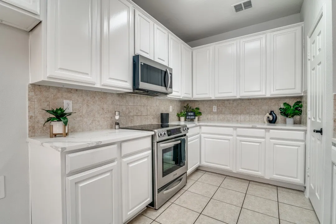 Kitchen featuring white shaker-style cabinetry, stainless steel appliances, light-toned tiled backsplash, quartz countertops, and tiled flooring
