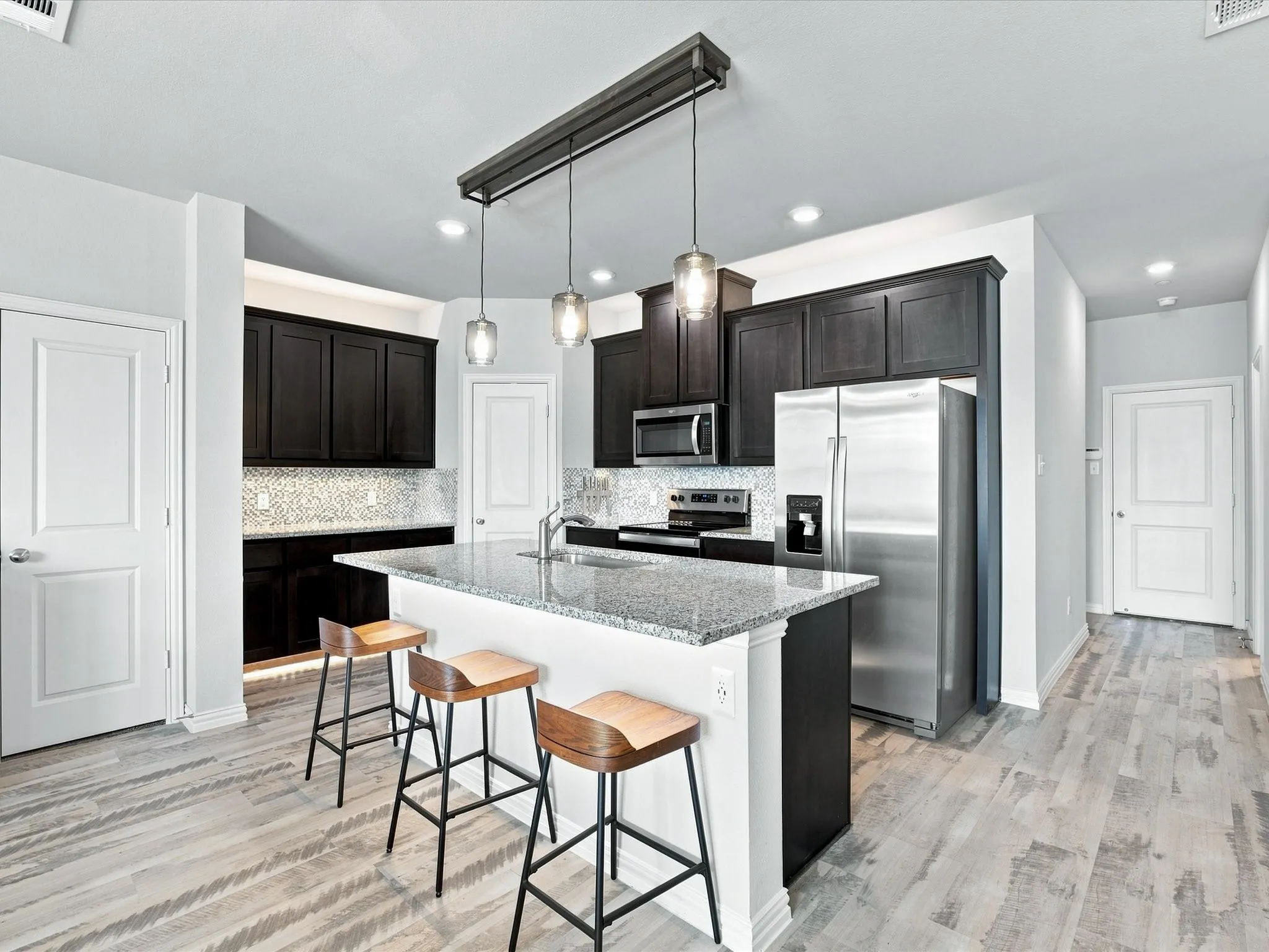 Kitchen featuring wood-finish flooring, dark wood-finish cabinetry, stainless steel appliances, a tile backsplash, and a large island with a stone countertop